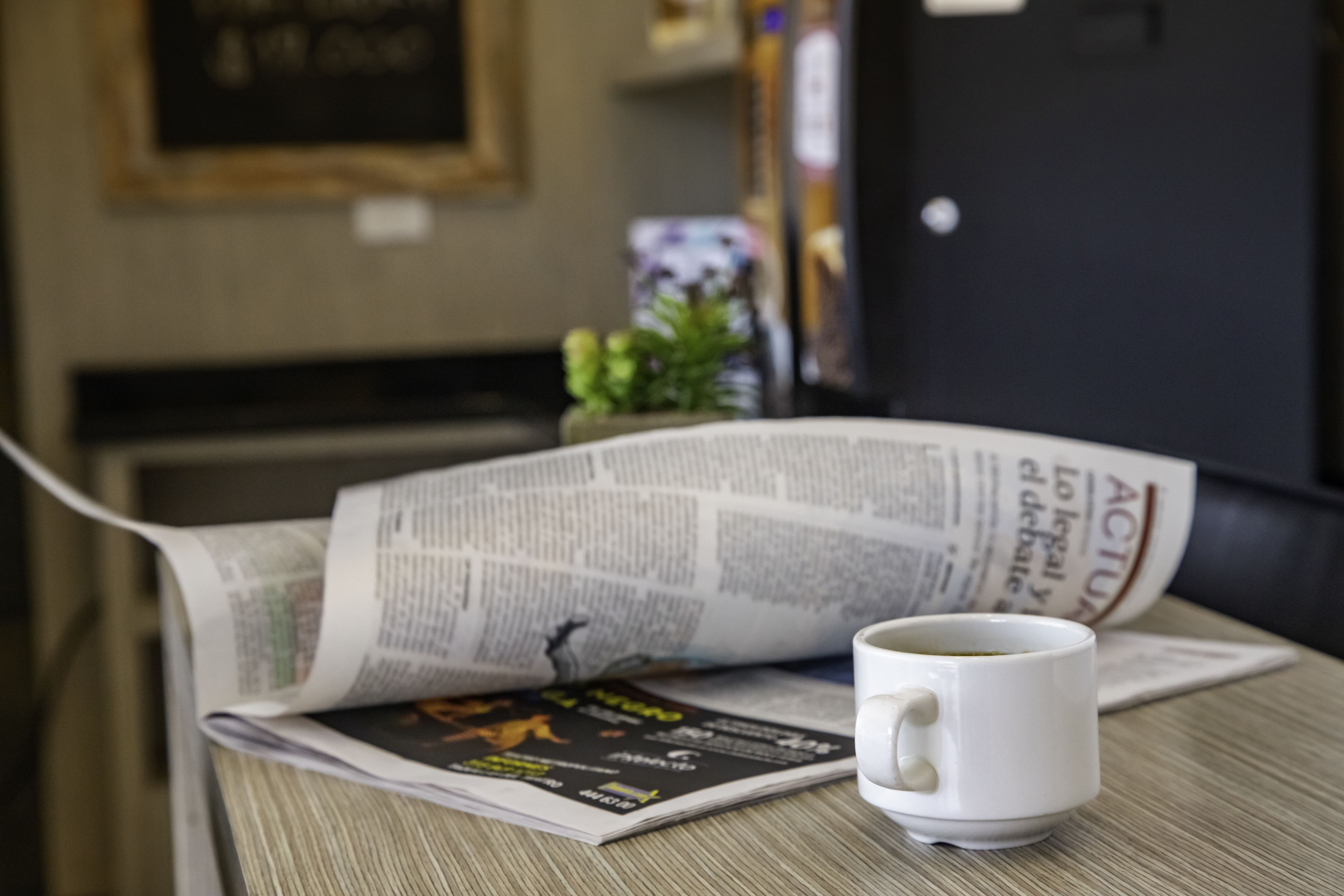 Um jornal, uma caneca branca e uma pequena planta sobre uma mesa de madeira.