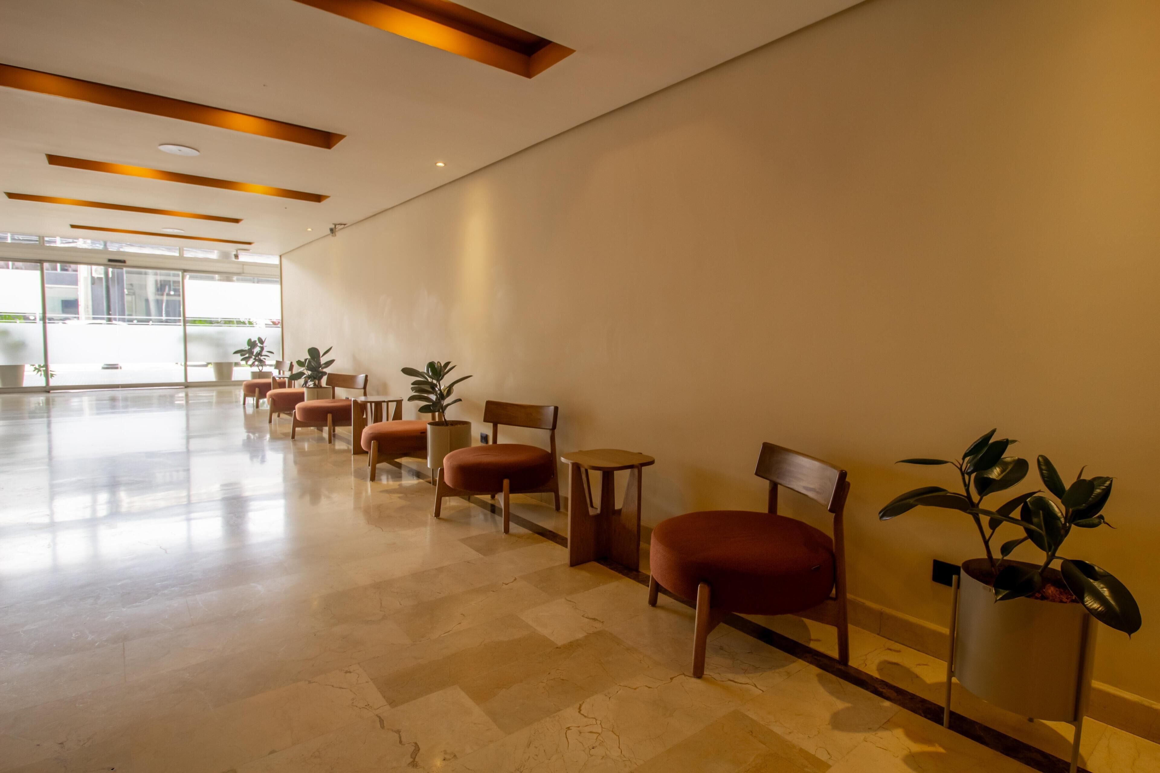 A spacious lobby with a long row of wooden chairs and tables, potted plants, and a large window providing a view of the outside.