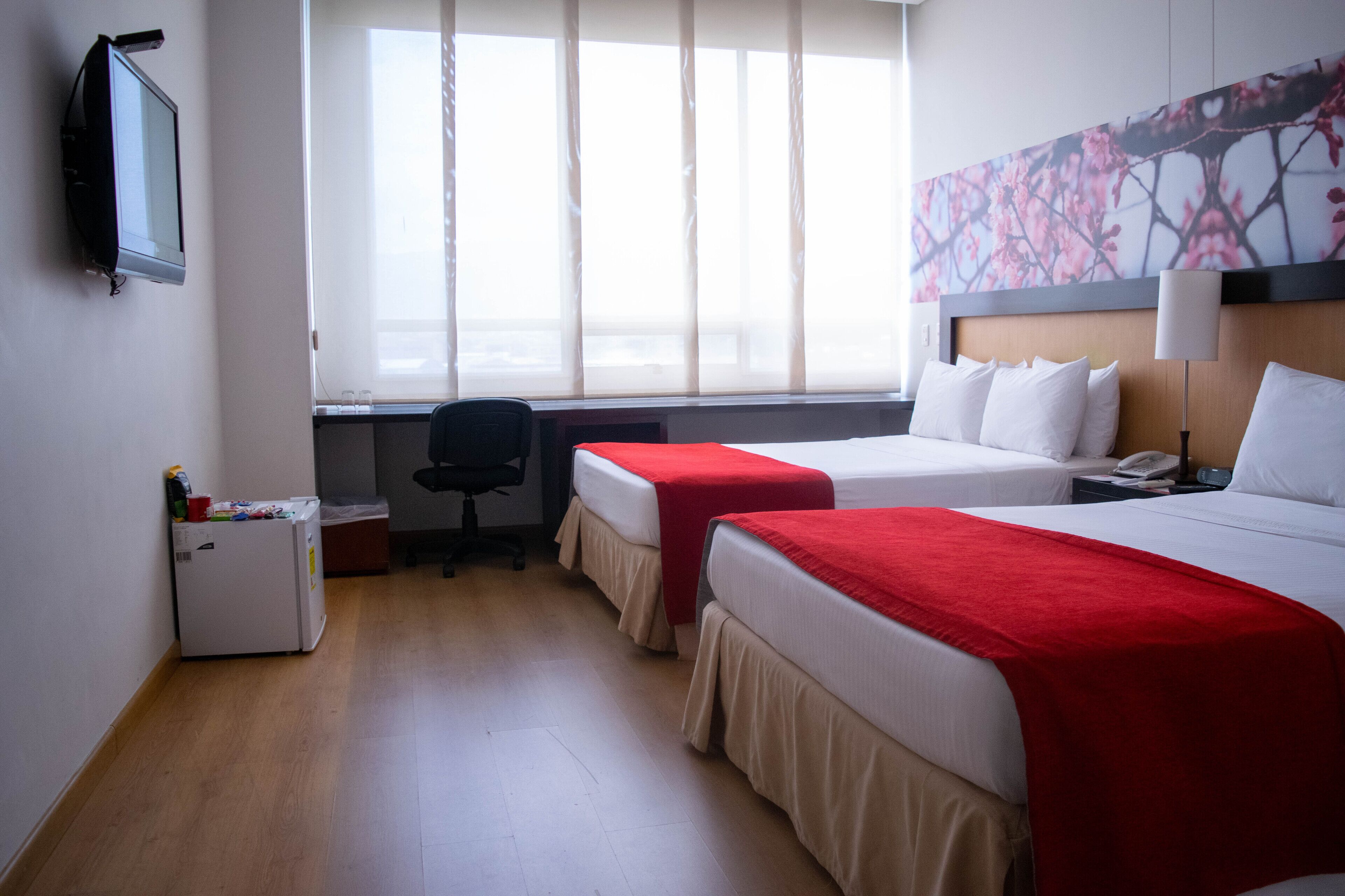 A hotel room with two beds, a desk, a refrigerator, and a television.