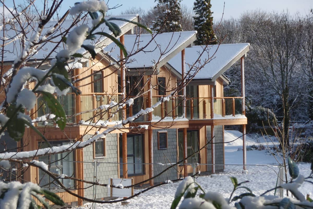 A modern two-story house with a balcony, surrounded by snow-covered trees and a clear sky.