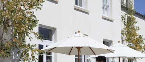 An outdoor seating area with tables and chairs, a waiter serving drinks, and a building with large windows and a stone wall in the foreground.