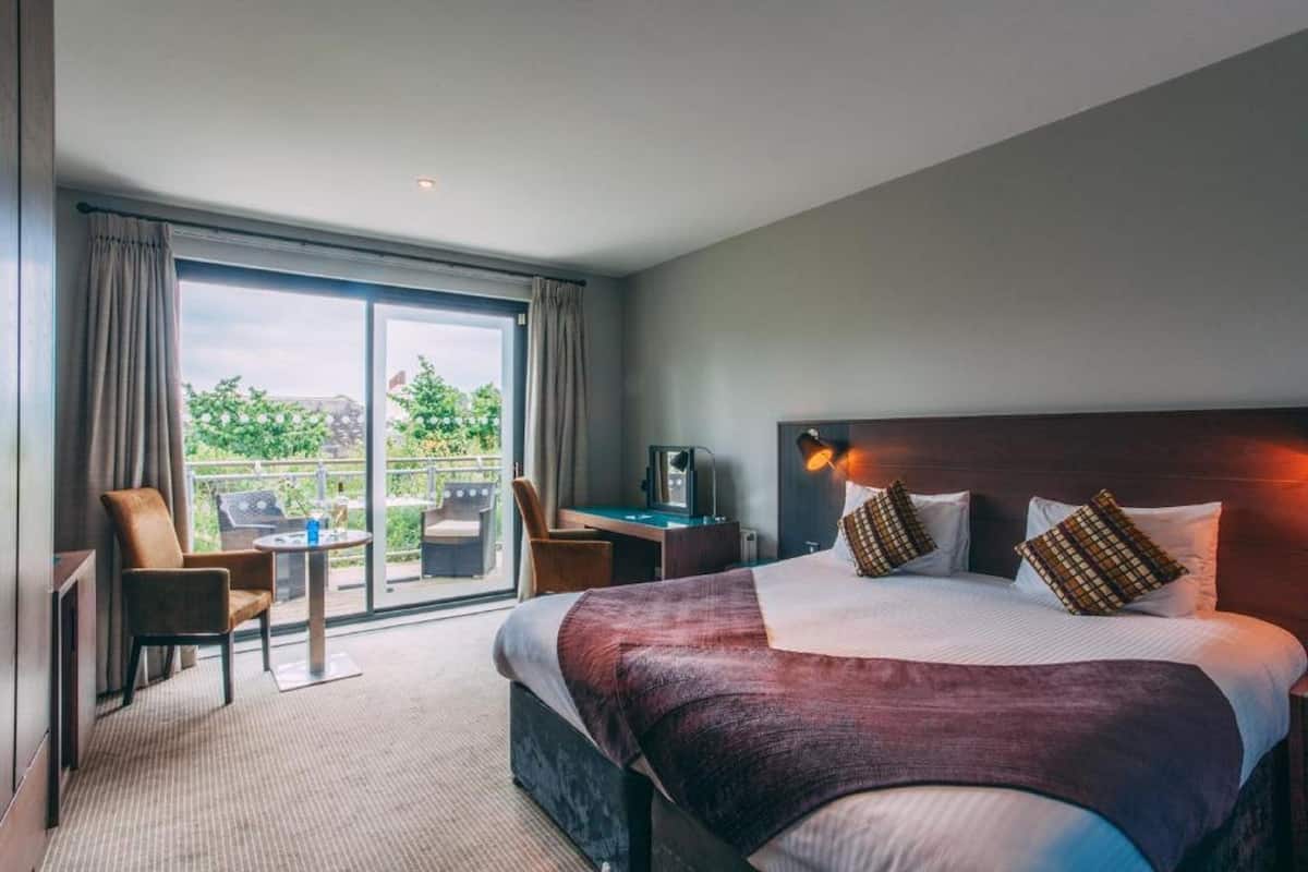 A hotel room with a large bed, a desk, a chair, and a balcony with a view of greenery.