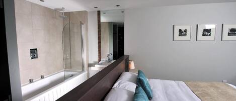 A modern hotel room with a large bed, a built-in shower, a large mirror, and a wall-mounted sink.
