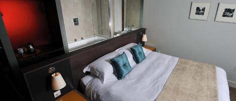 Una habitación de hotel moderna con una cama grande, una mesita de noche con lámpara, un espejo y dos cuadros en la pared.