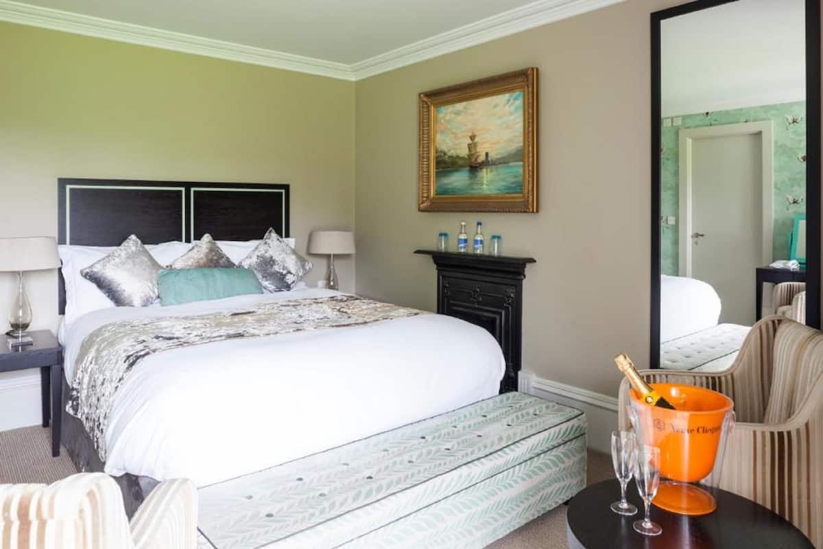 A hotel room with a large bed, a nightstand with a lamp, a painting on the wall, a chair, and a table with a champagne bucket and glasses.