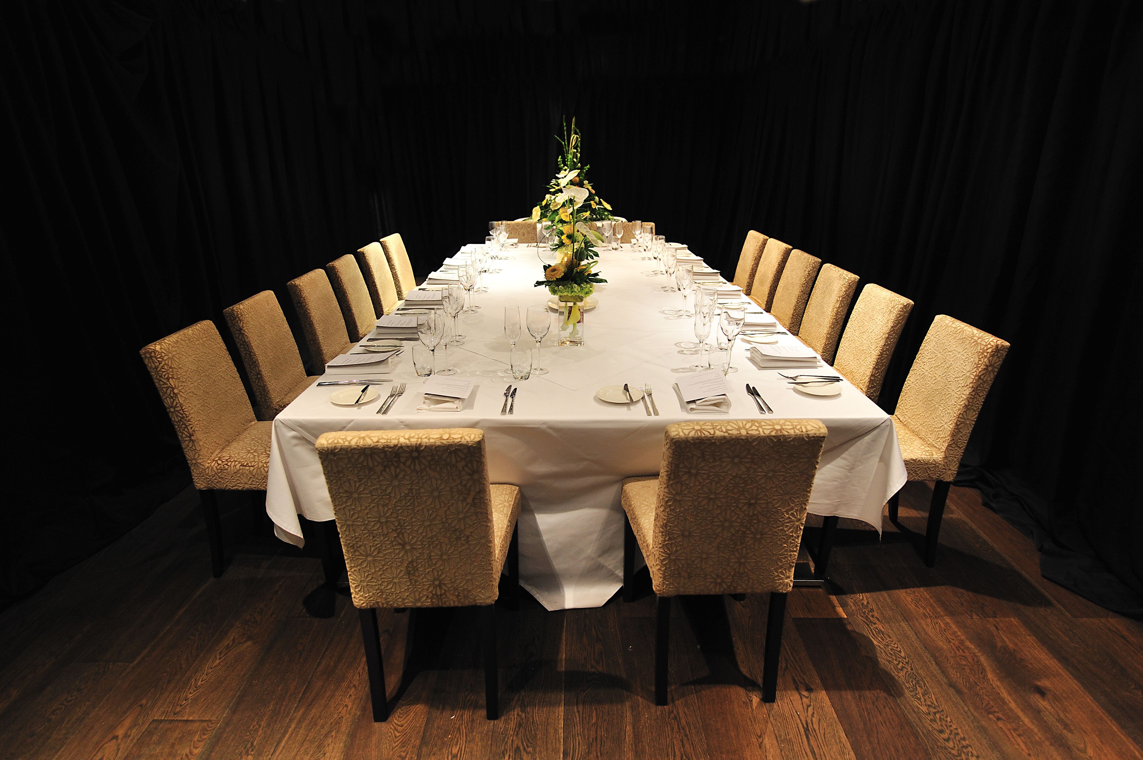 A long dining table set for a formal meal with chairs, a centerpiece, and a dark curtain backdrop.
