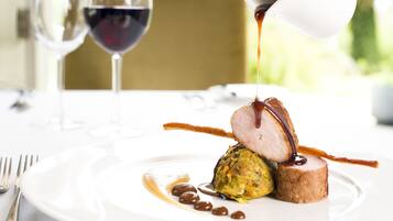 A plated dish with a slice of meat, a dollop of sauce, and a garnish, with a wine glass in the background.