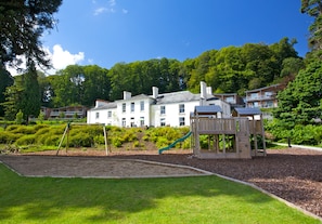A playground with a slide and swings, surrounded by a well-maintained lawn and a large building in the background.