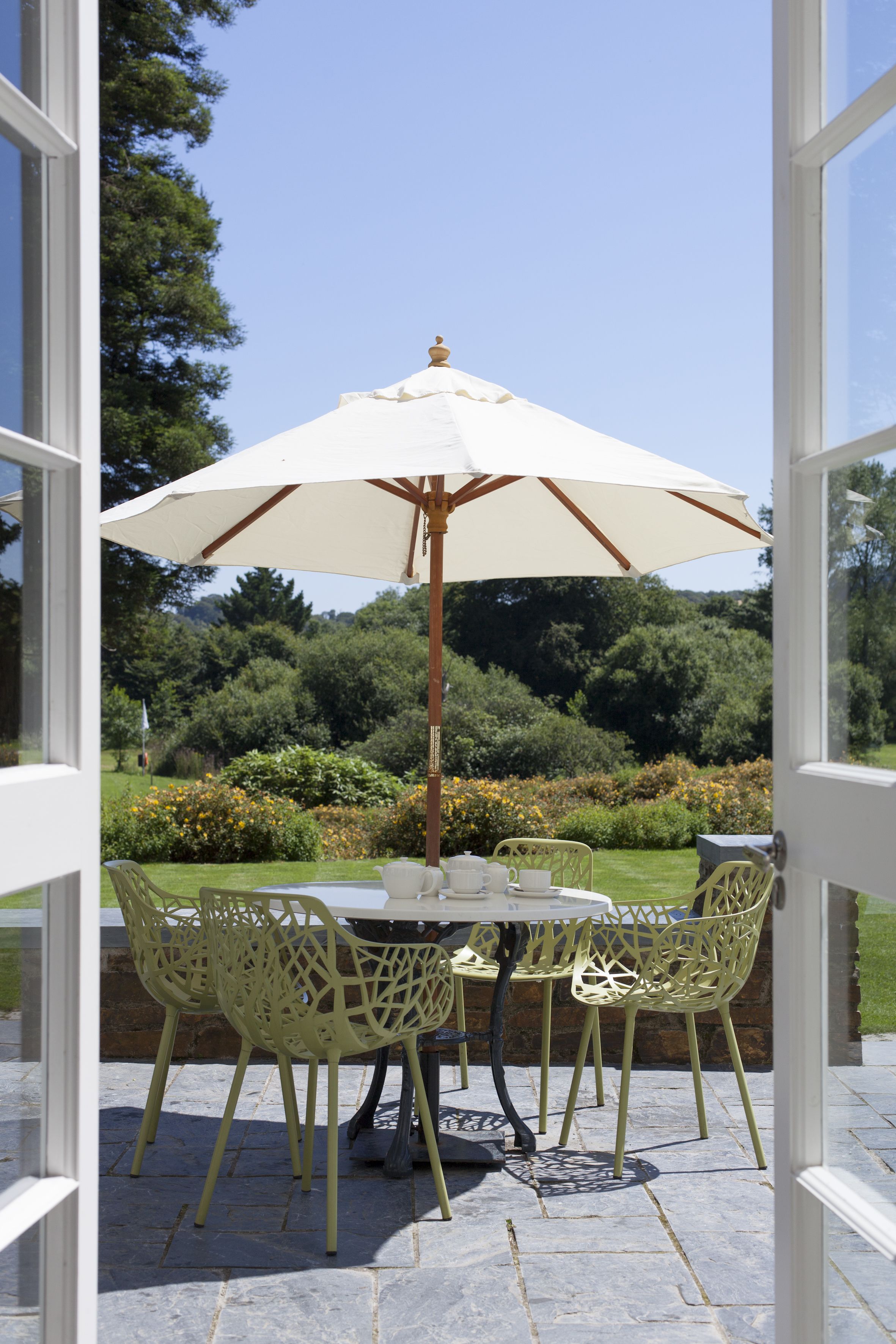 An outdoor patio with a table and chairs, a white umbrella, and a view of a garden.