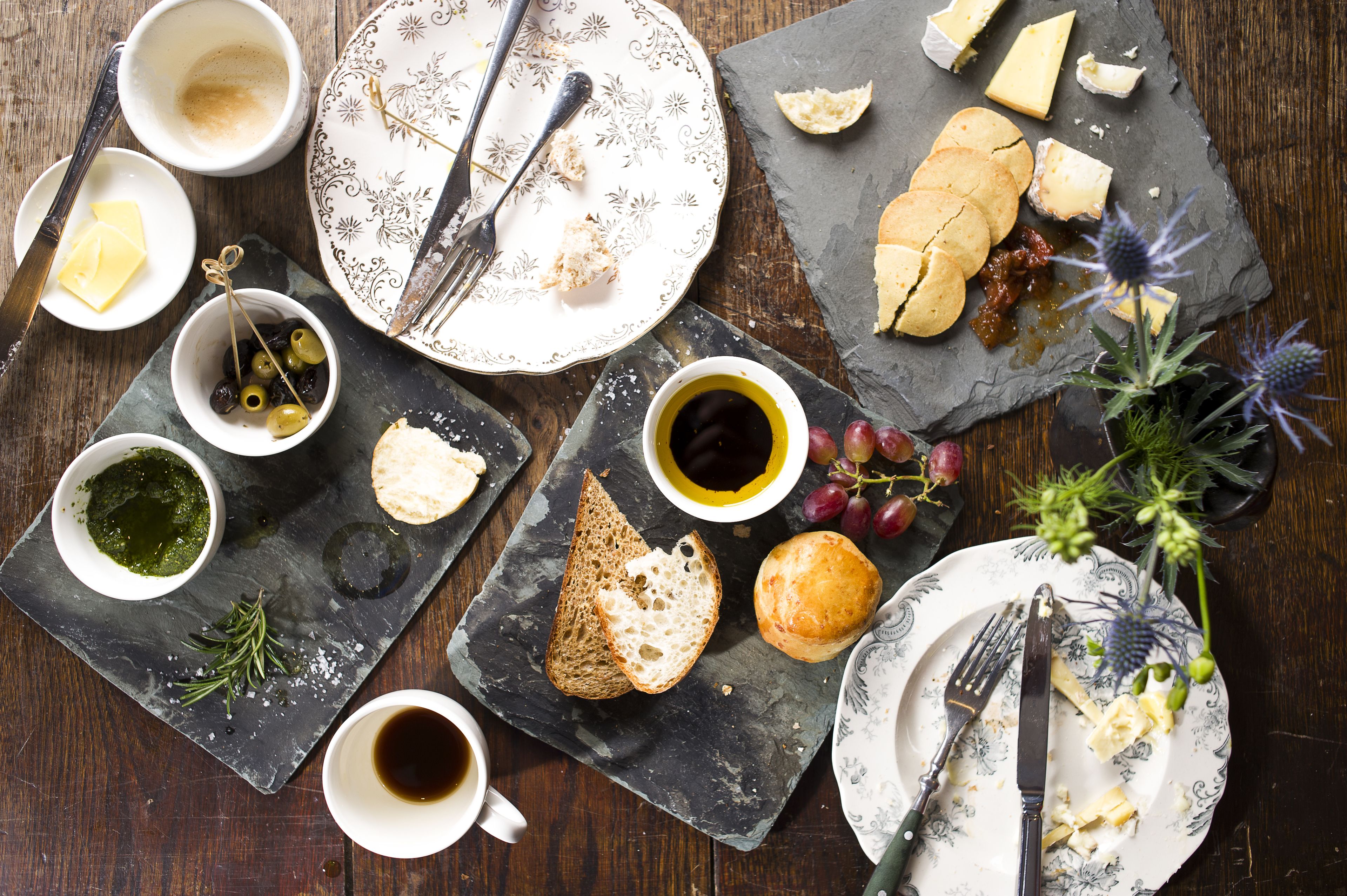 Uma mesa com diversos alimentos, incluindo queijo, pão e azeitonas, além de xícaras de café e um prato com garfo e faca.