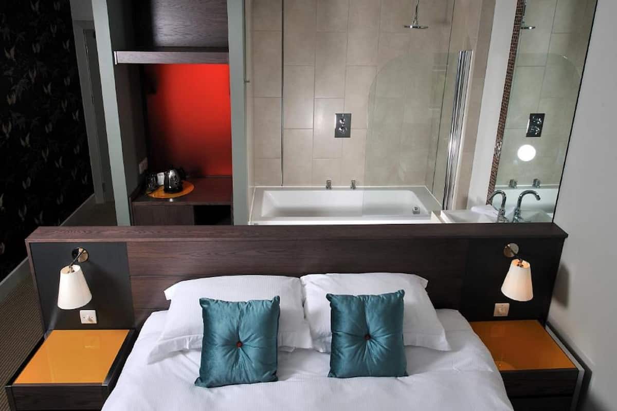 A hotel room with a bed, two bedside lamps, a nightstand, a red-lit shelf, a bathtub, and a large mirror.