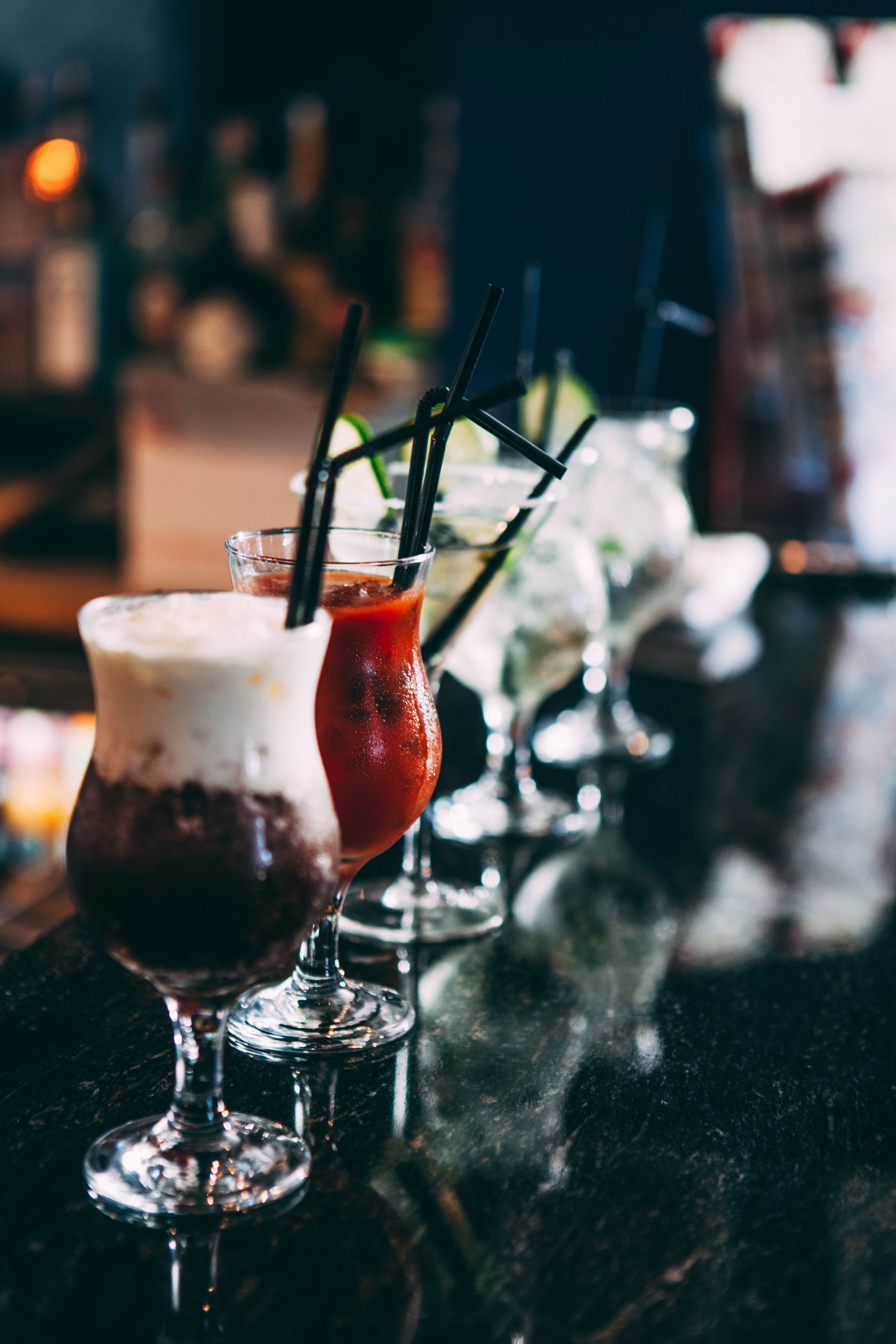A bar setting with various glasses of drinks, including a layered cocktail and a dark beverage, on a reflective surface.