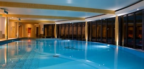 Una amplia piscina interior con agua cristalina y azul, rodeada por paredes de cristal y vigas de madera en el techo.