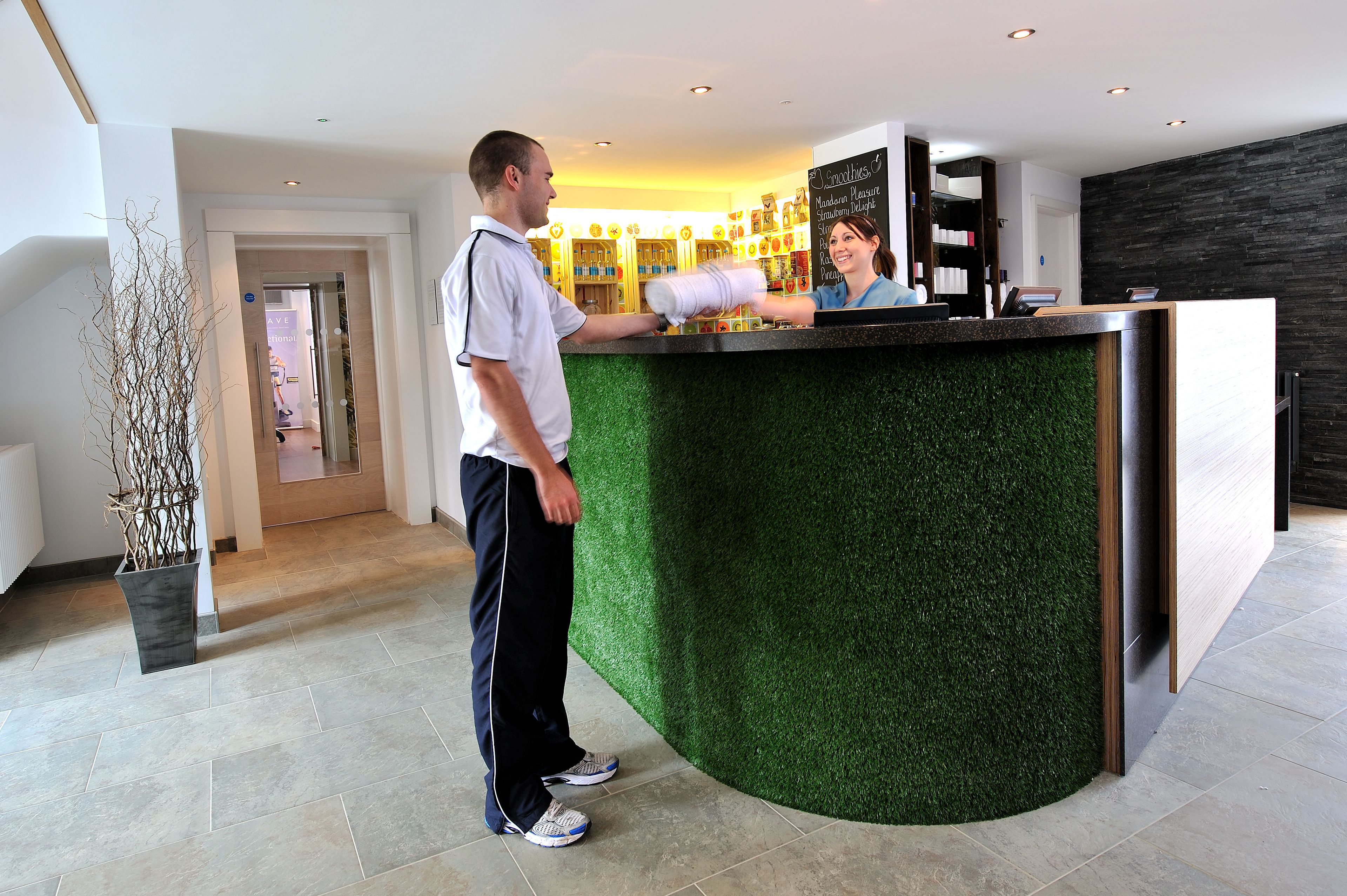 Un comptoir d’accueil avec un plateau recouvert d’un gazon artificiel vert, un mur blanc sur lequel est affiché un menu sur un tableau noir, et une plante en pot placée à gauche.