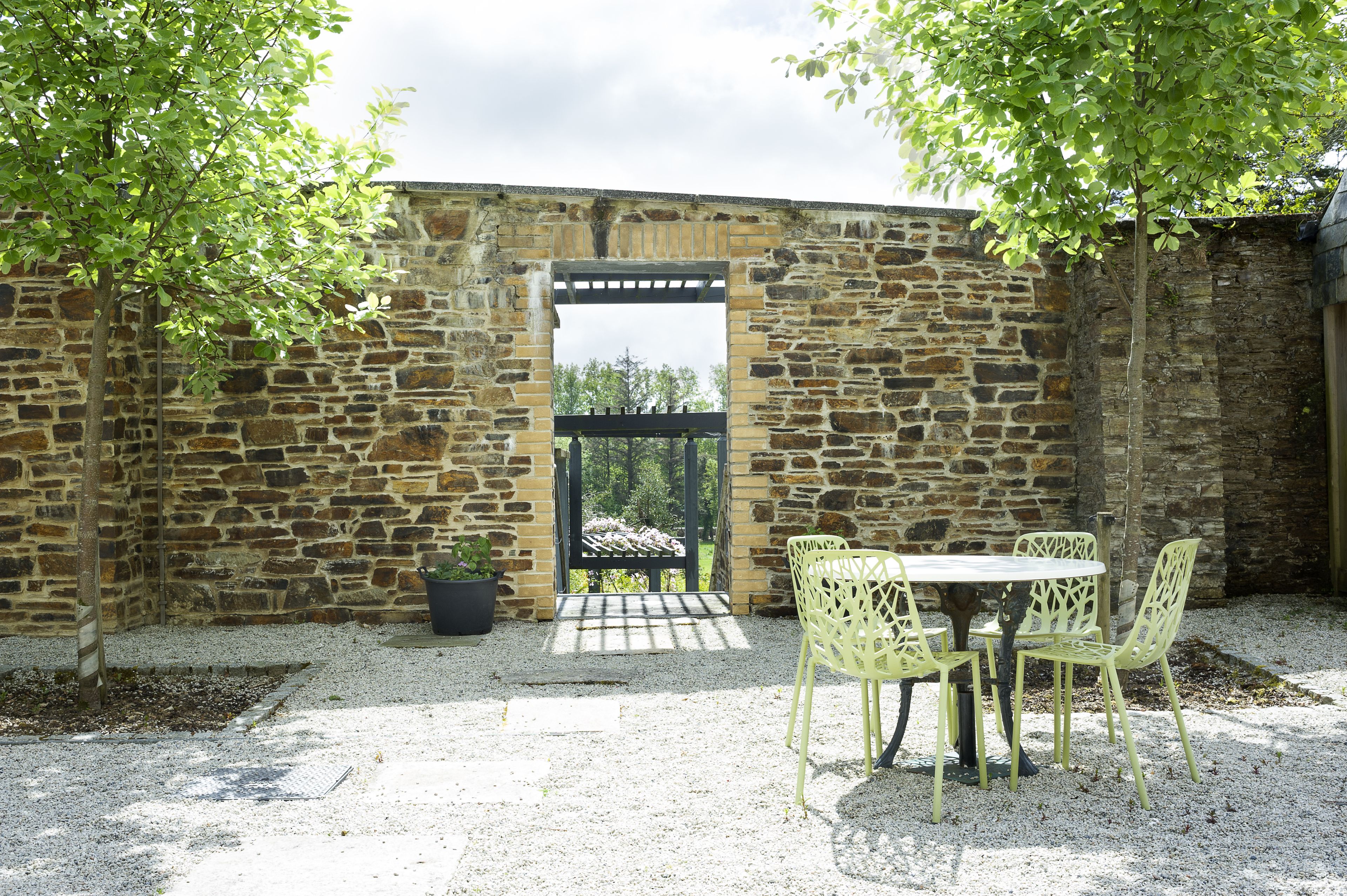 Un muro de piedra con un arco de pérgola de madera, un patio de grava con una mesa redonda y sillas, y una planta en maceta.