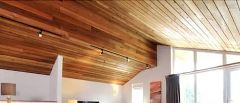 A modern living room with a wooden ceiling, a flat-screen TV, a sofa, a coffee table, and a large window.