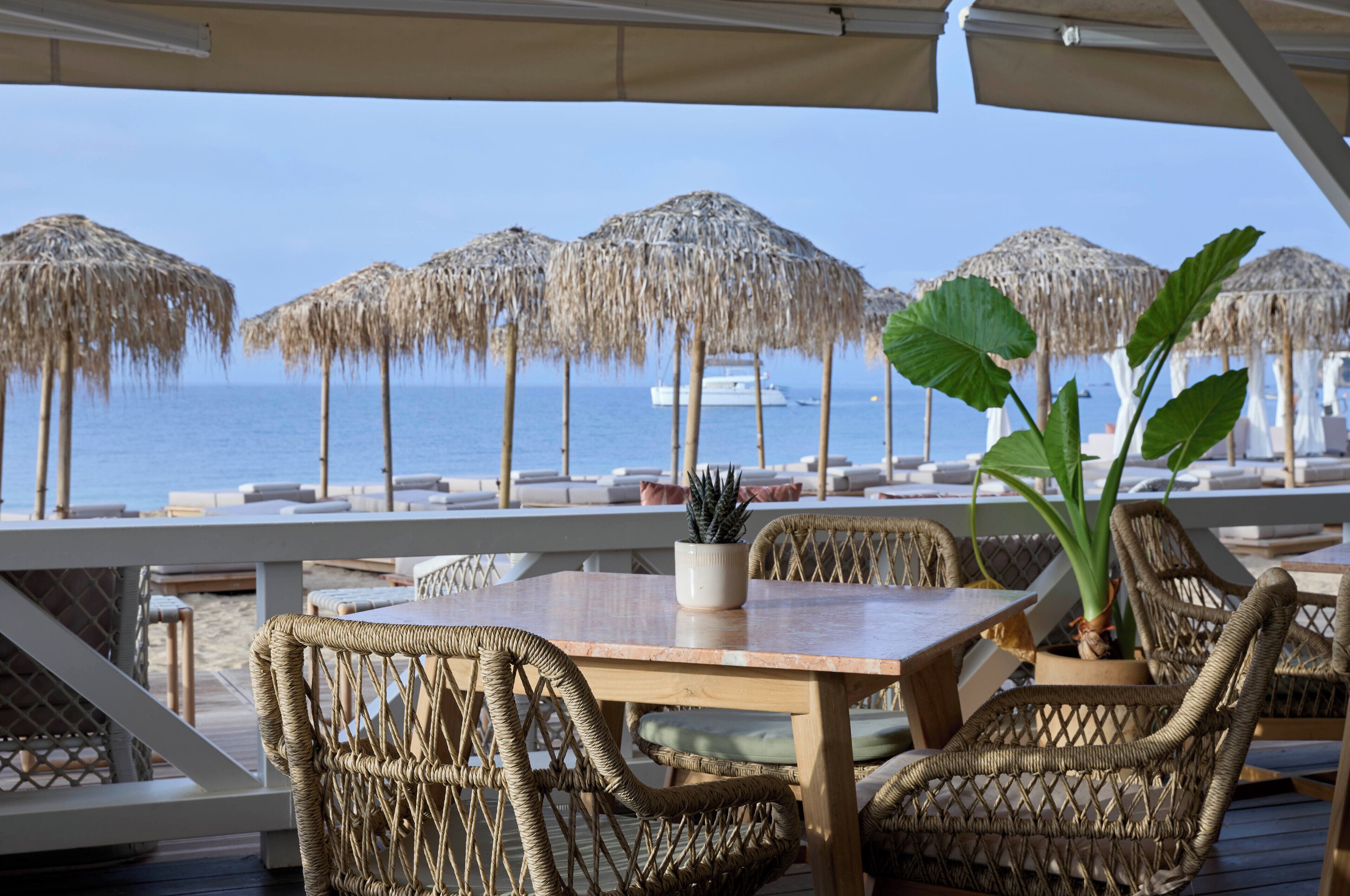 A beachside patio with wicker furniture, a wooden table, and thatched umbrellas.