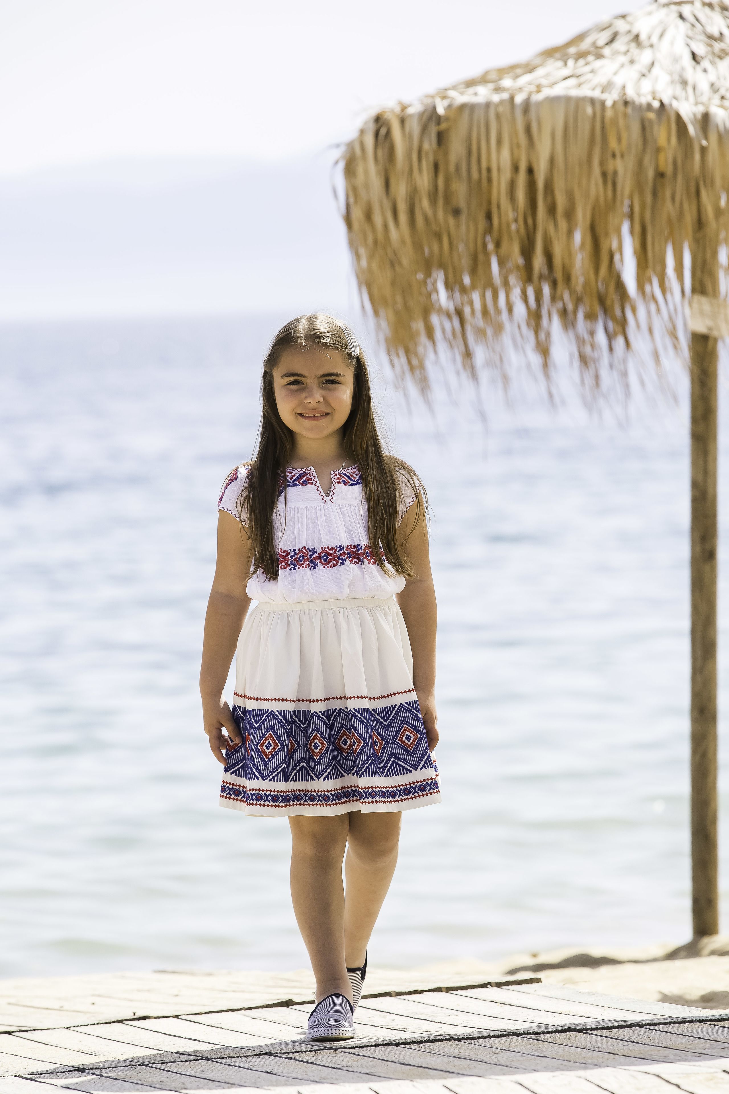 A young girl in a patterned dress walking on a wooden deck by the sea.