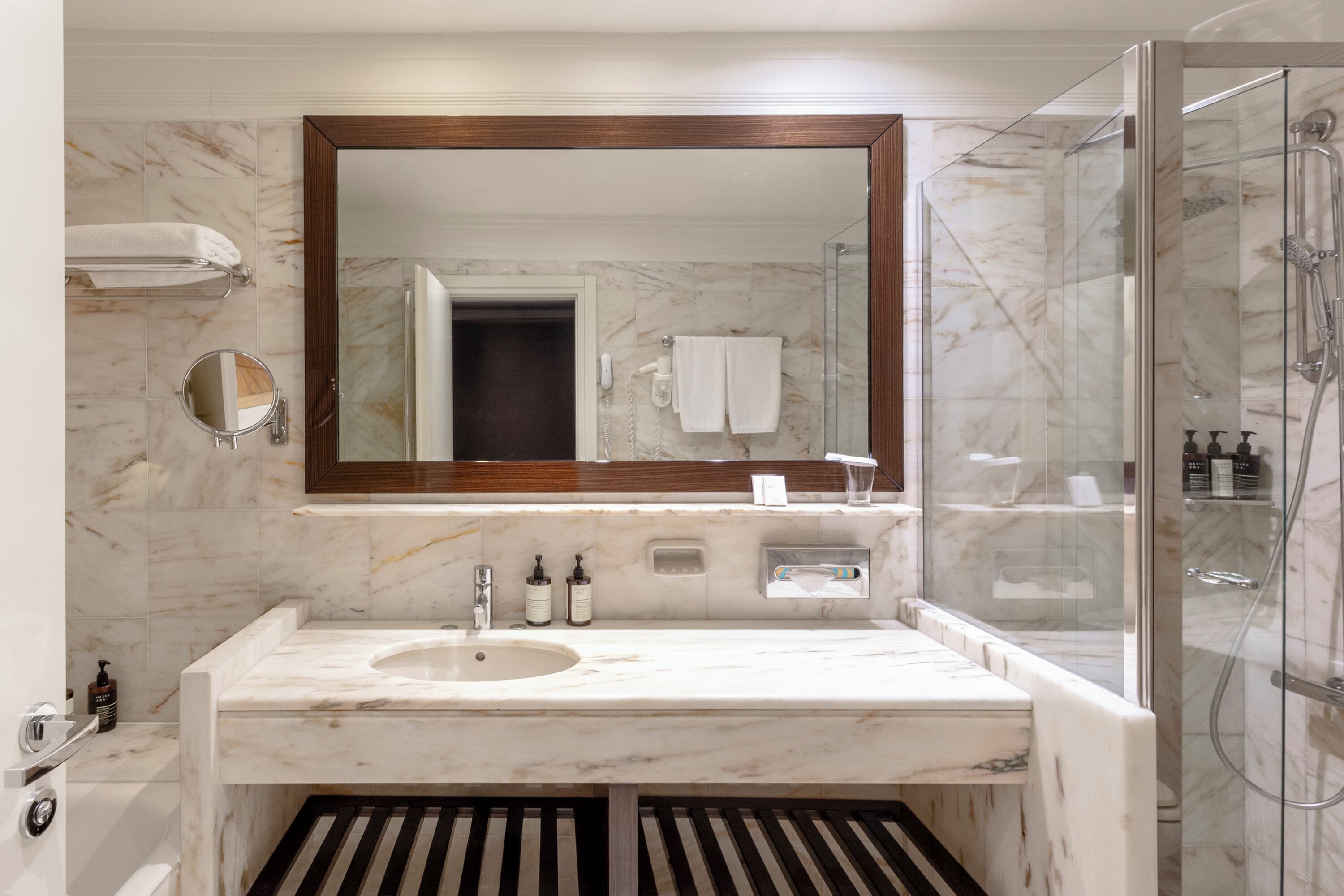 Un baño moderno con lavamanos de mármol, un espejo grande, ducha con mampara de vidrio y un perchero para toallas.