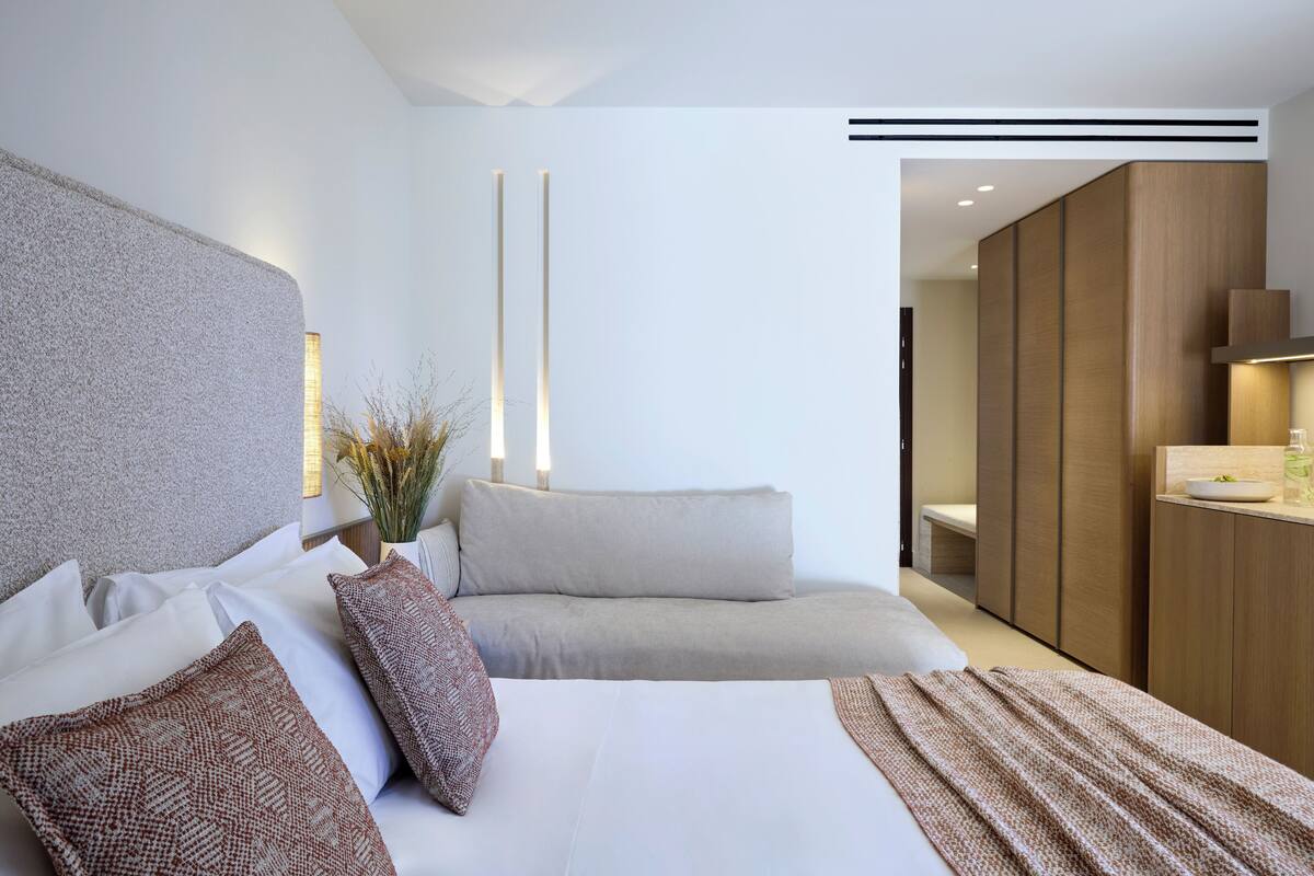 A modern hotel room with a large bed, a sofa, a wooden wardrobe, and a bathroom visible through an open door.