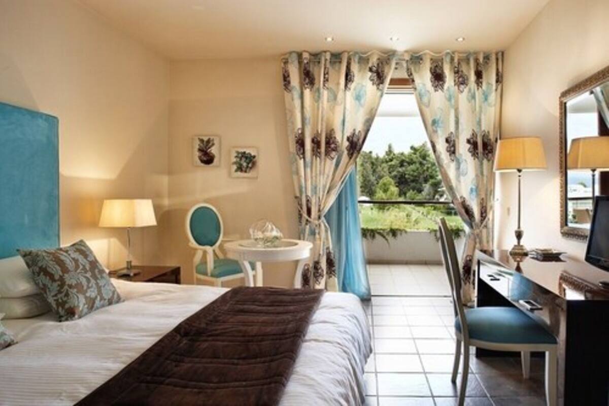 A hotel room with a bed, a desk with a chair, a small table with a lamp, and a view of greenery outside.