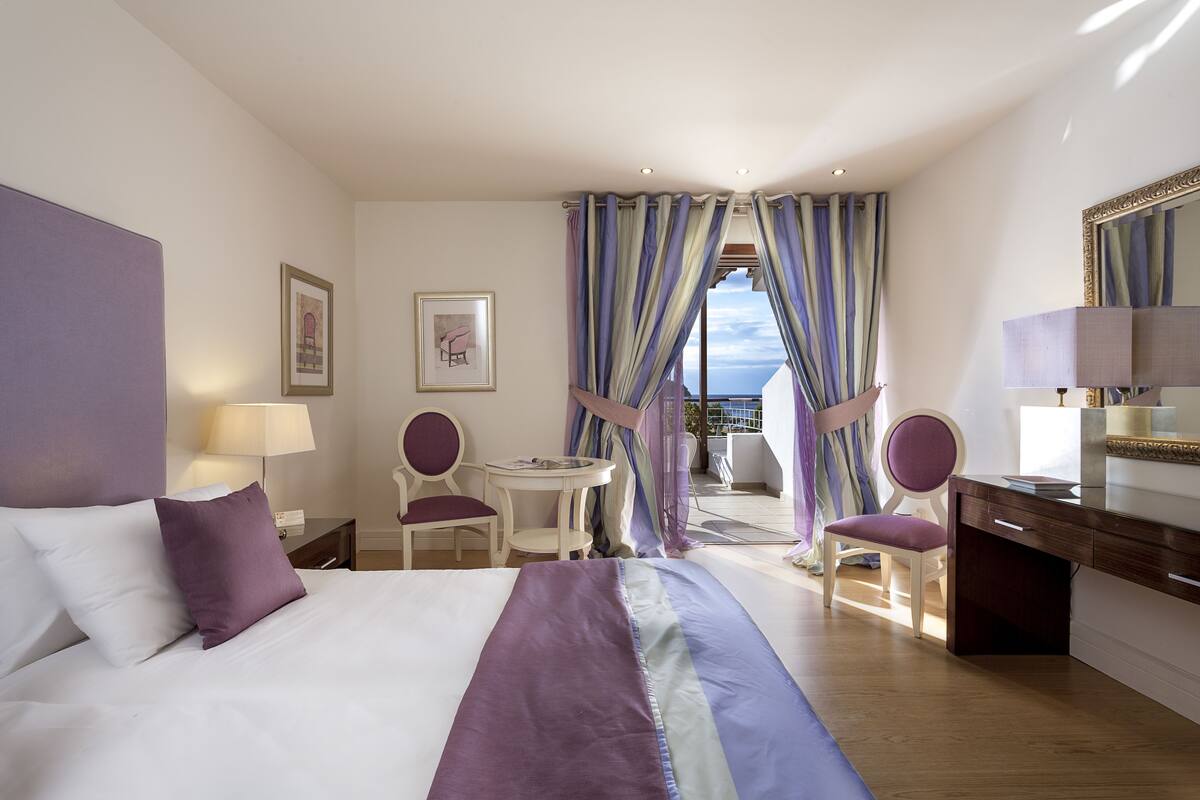 A hotel room with a bed, a desk, two chairs, a small table, and a balcony with a view.