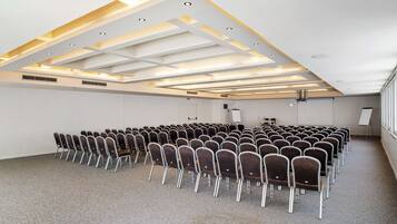 Un salón de conferencias con filas de sillas, un techo blanco con iluminación empotrada y ventanas amplias en un lateral.