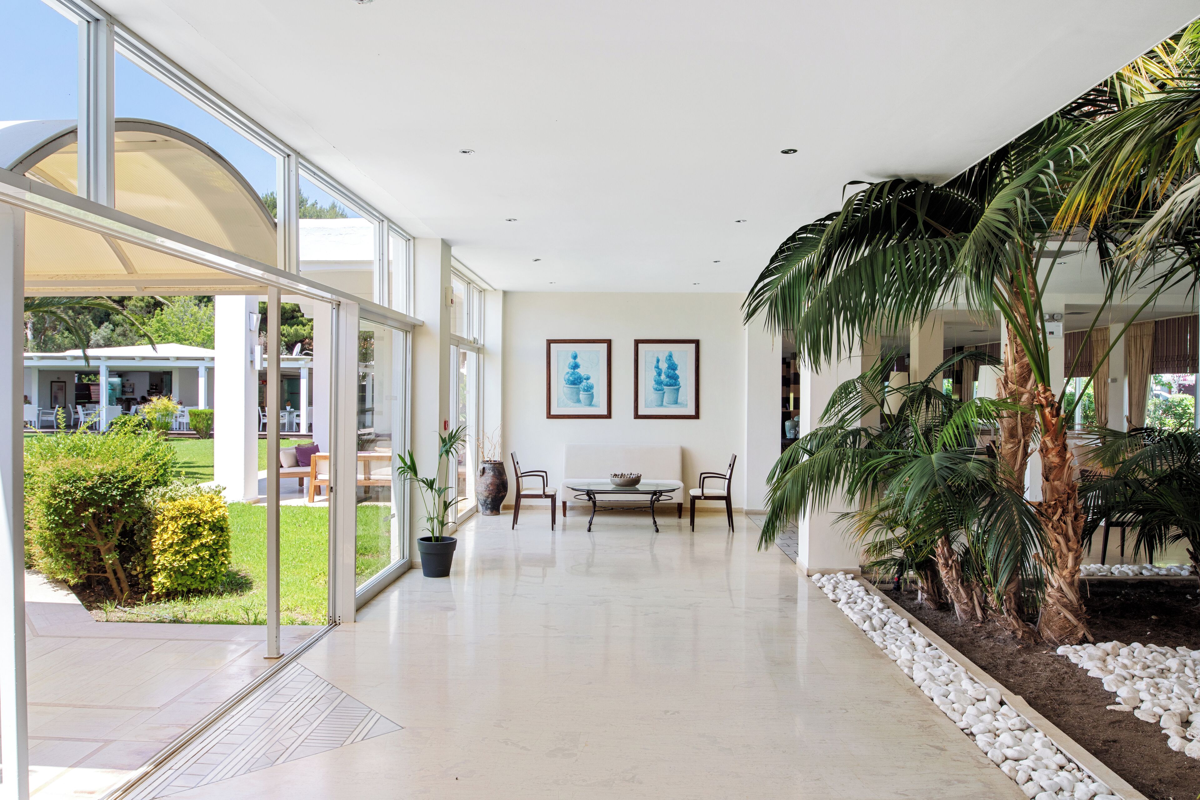 Un espacio interior amplio con pared de vidrio que ofrece vista al exterior, zona de comedor con mesa y sillas, y pequeño jardín con plantas y una palmera.