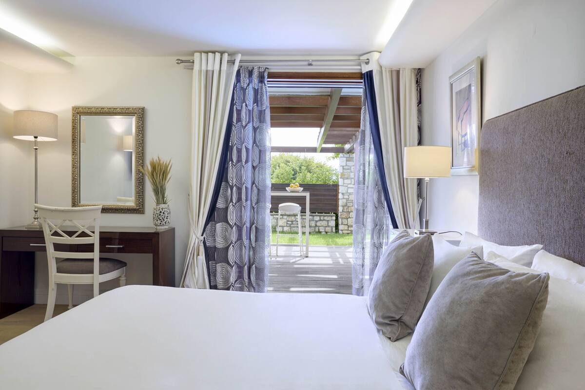 A bedroom with a bed, a desk, a chair, a mirror, and a view of an outdoor area.