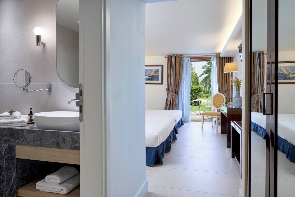 A hotel room with a large round mirror, a white sink, a bed with blue bedding, a desk with a lamp, and a view of palm trees through the window.