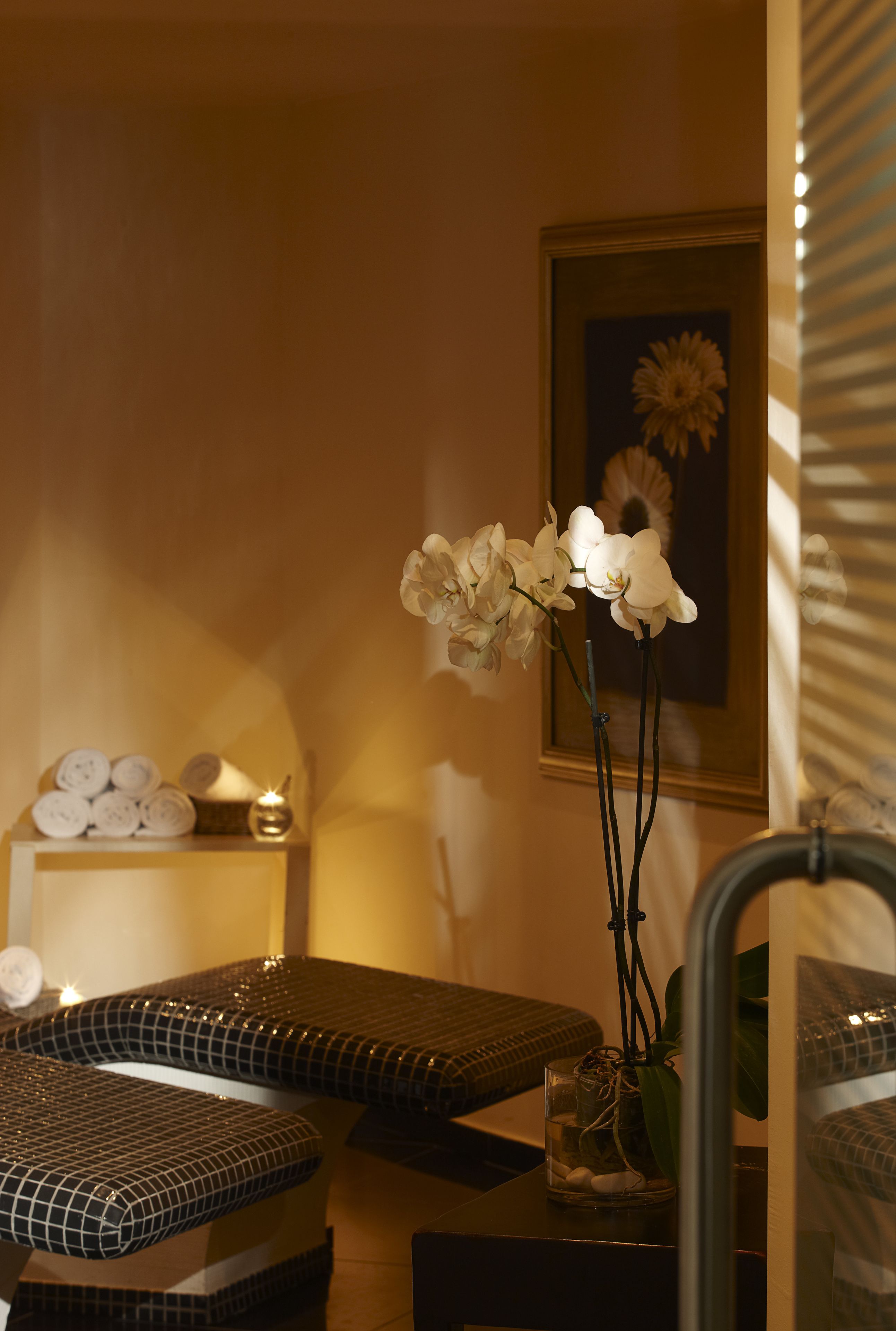 Une salle de spa équipée d’une table de massage, d’une petite étagère avec des serviettes et d’un vase contenant des fleurs blanches.