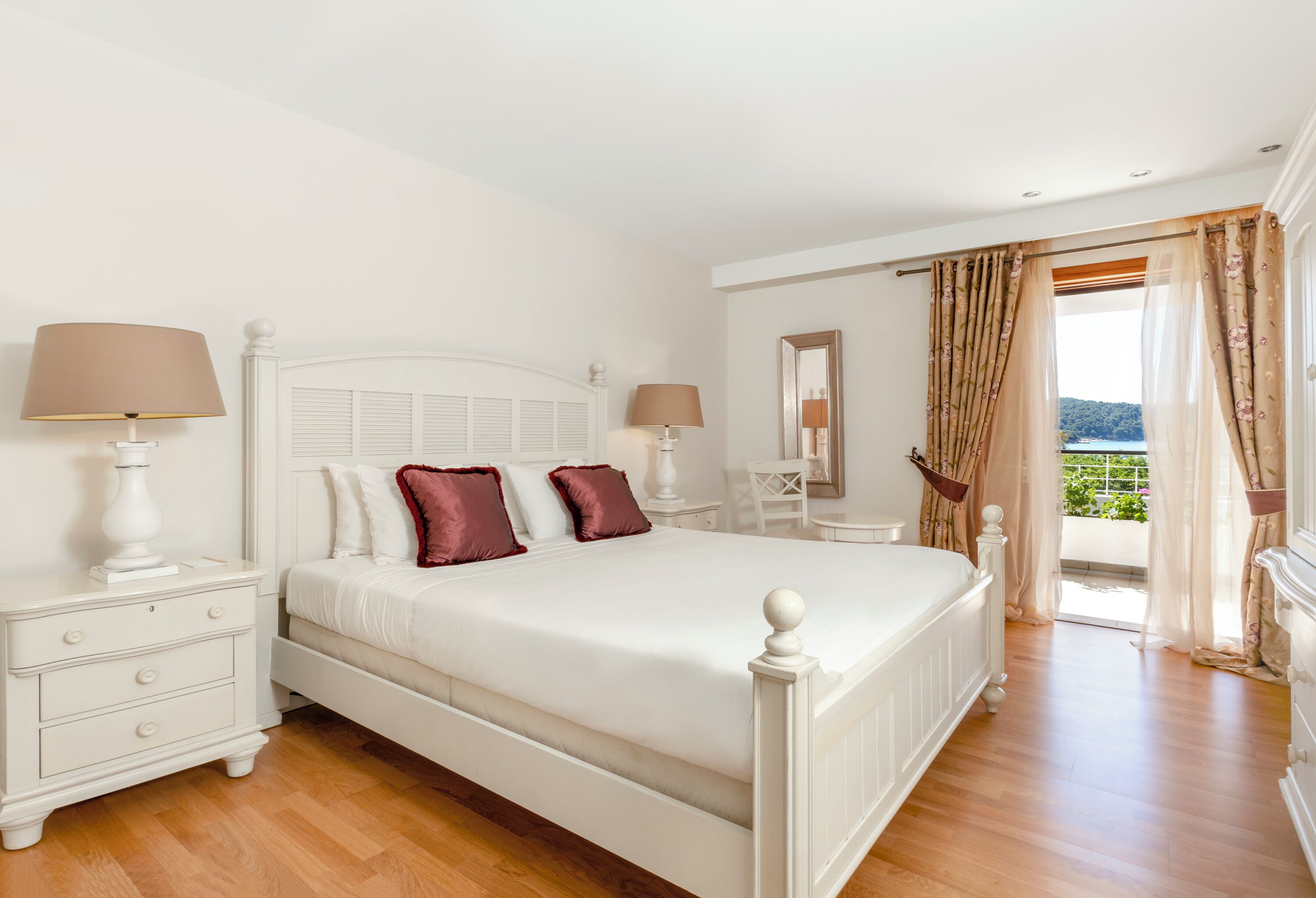 A bedroom with a bed, nightstands, a chair, a table, and a view through a window.