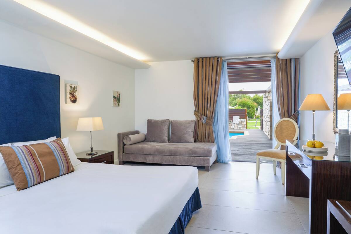 A hotel room with a bed, a sofa, a desk with a chair, a lamp, and a view of the outside.