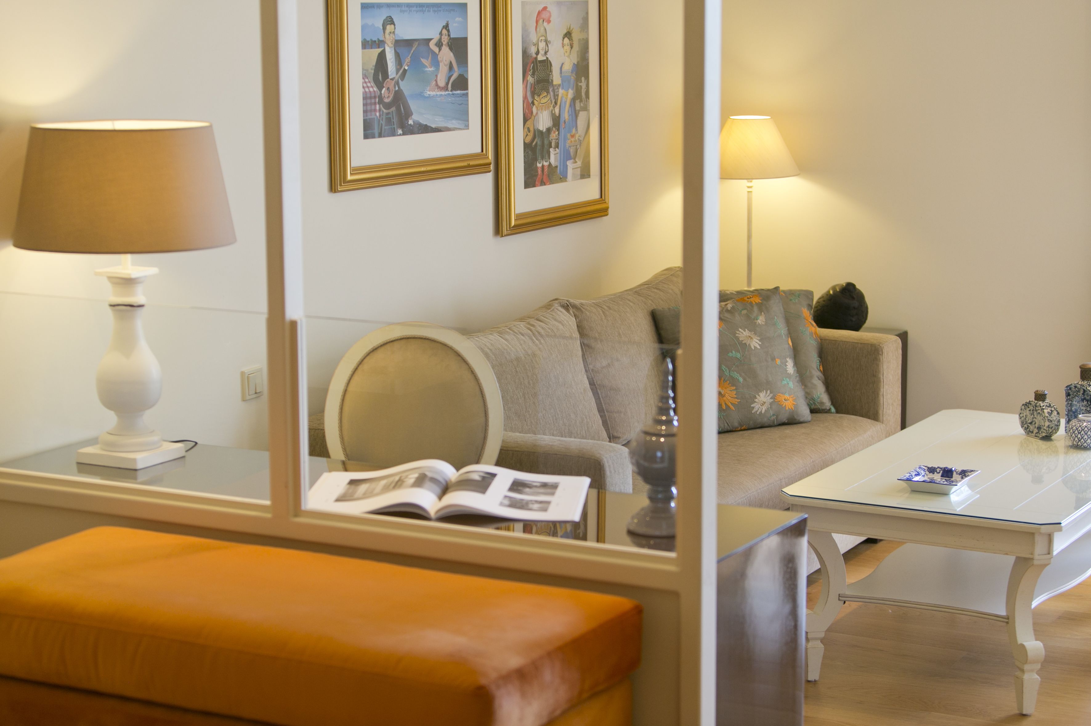 A modern living room with a sofa, a glass coffee table, and a lamp. There are framed pictures on the wall and a book on the table.