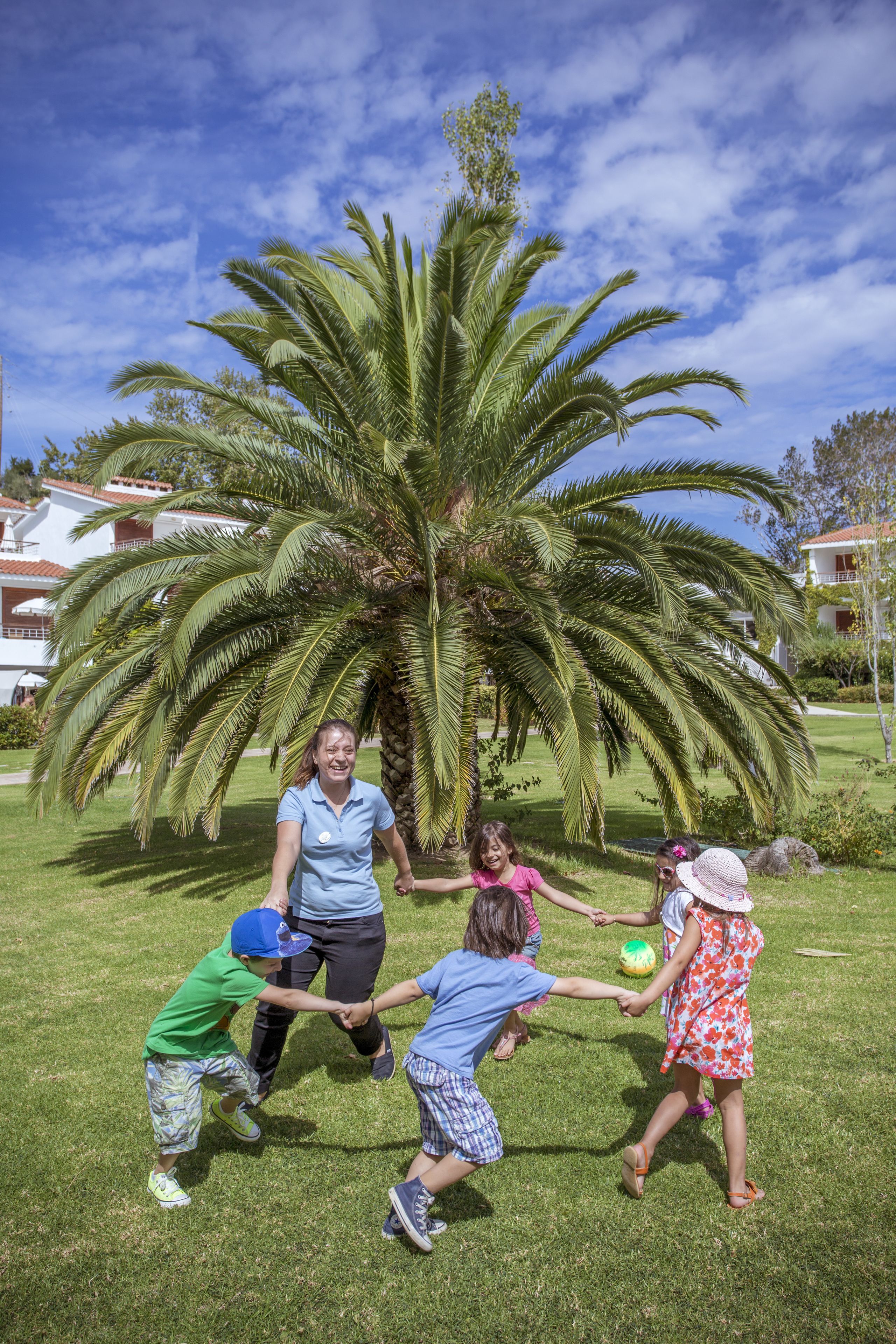 A group of children playing with a ball in a grassy area with a large palm tree and houses in the background.