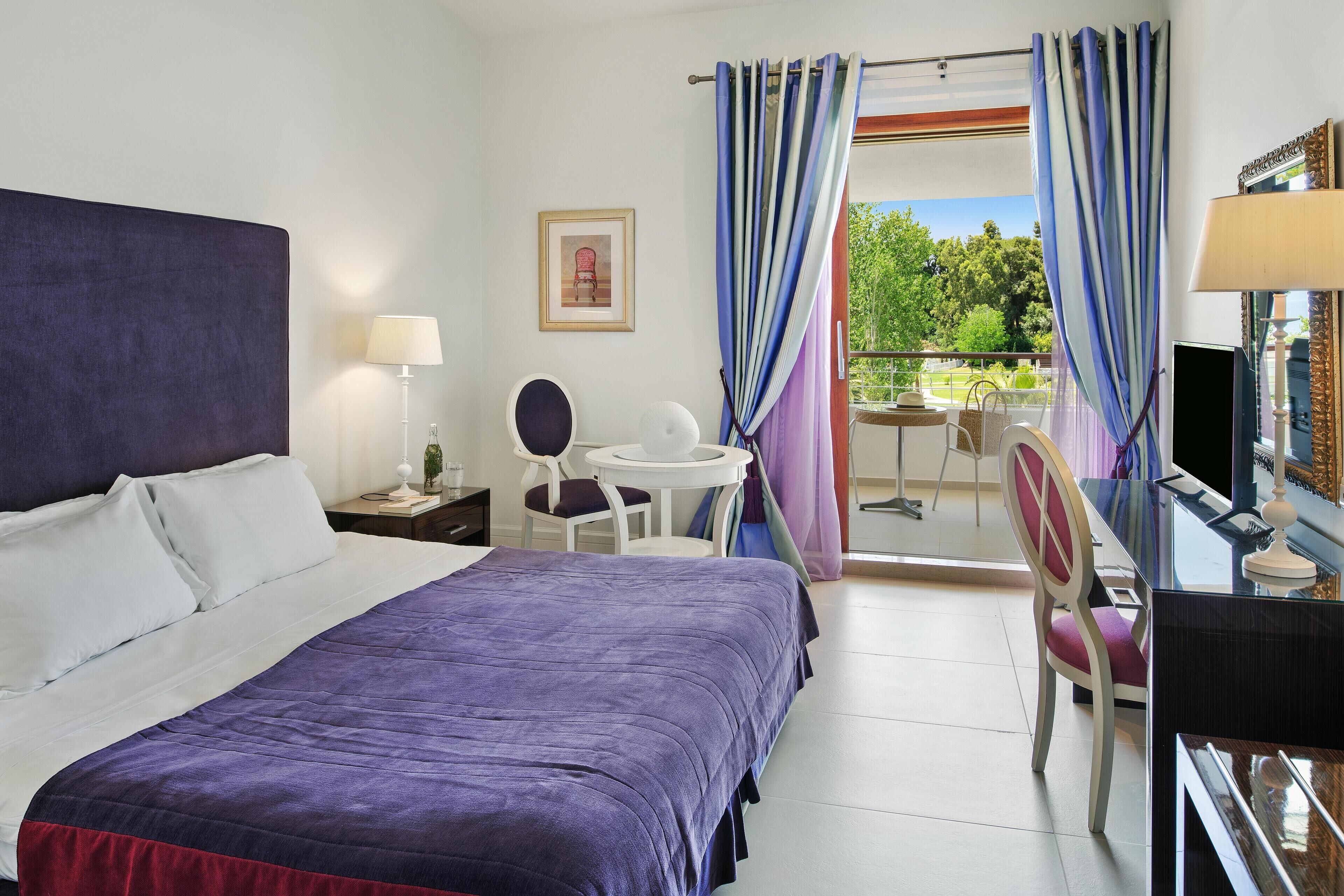 A hotel room with a bed, a desk with a chair, a small table with a chair, and a balcony with curtains.
