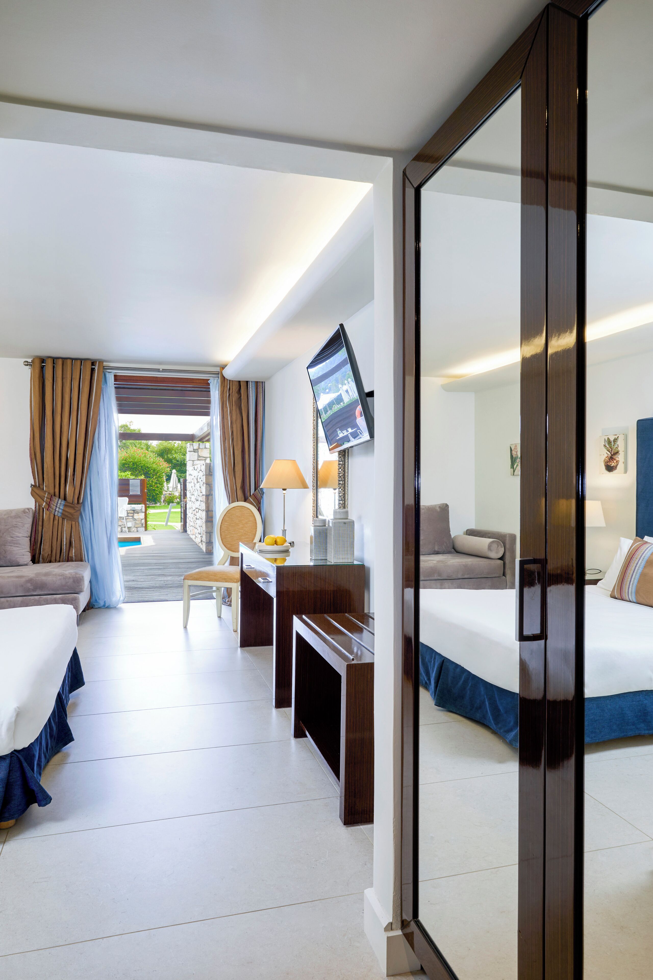 A hotel room with two beds, a desk, a chair, a TV, and a view of the outdoors.