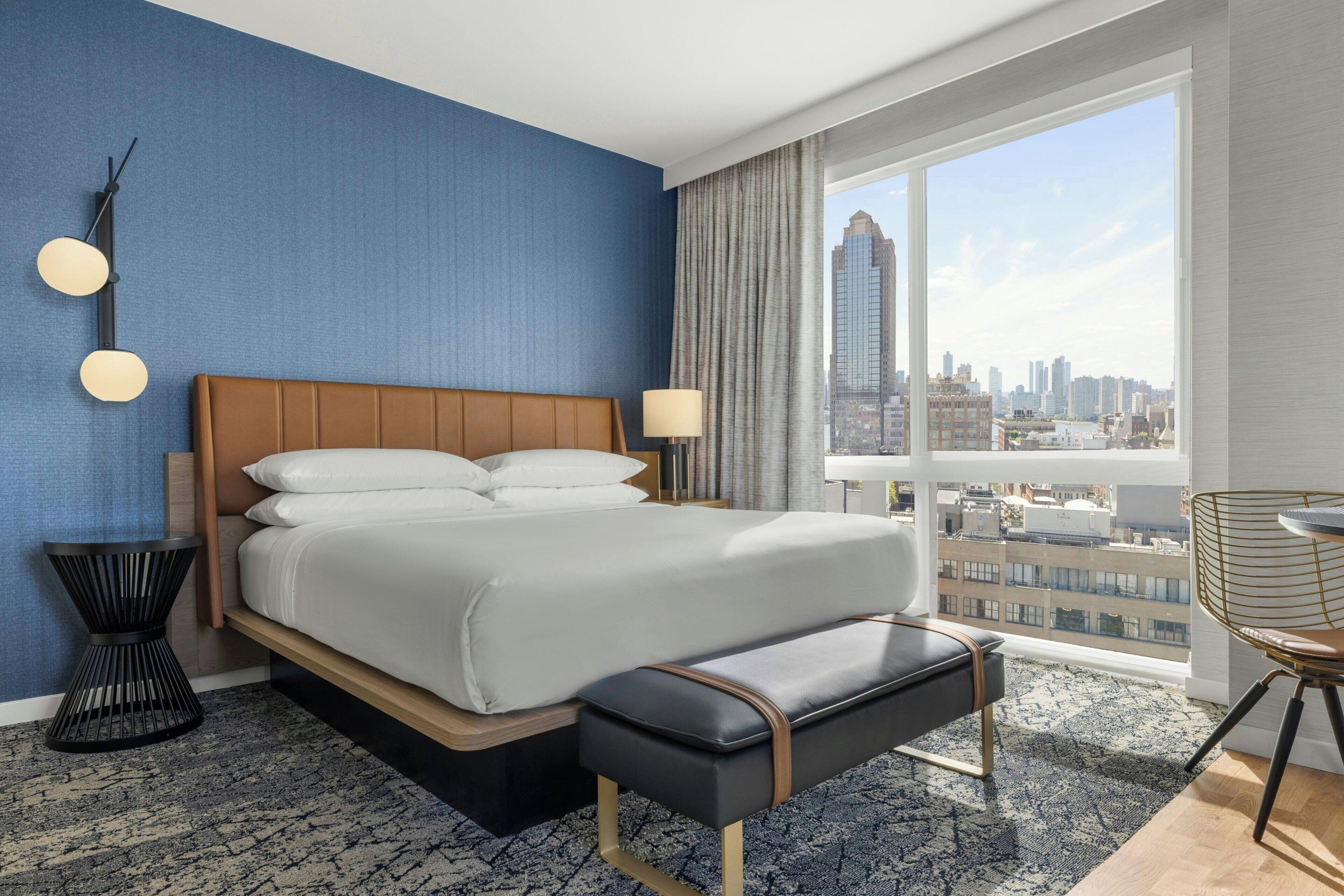 Une chambre d’hôtel avec un grand lit, une vue sur le paysage urbain, un fauteuil moderne et une petite table d’appoint.