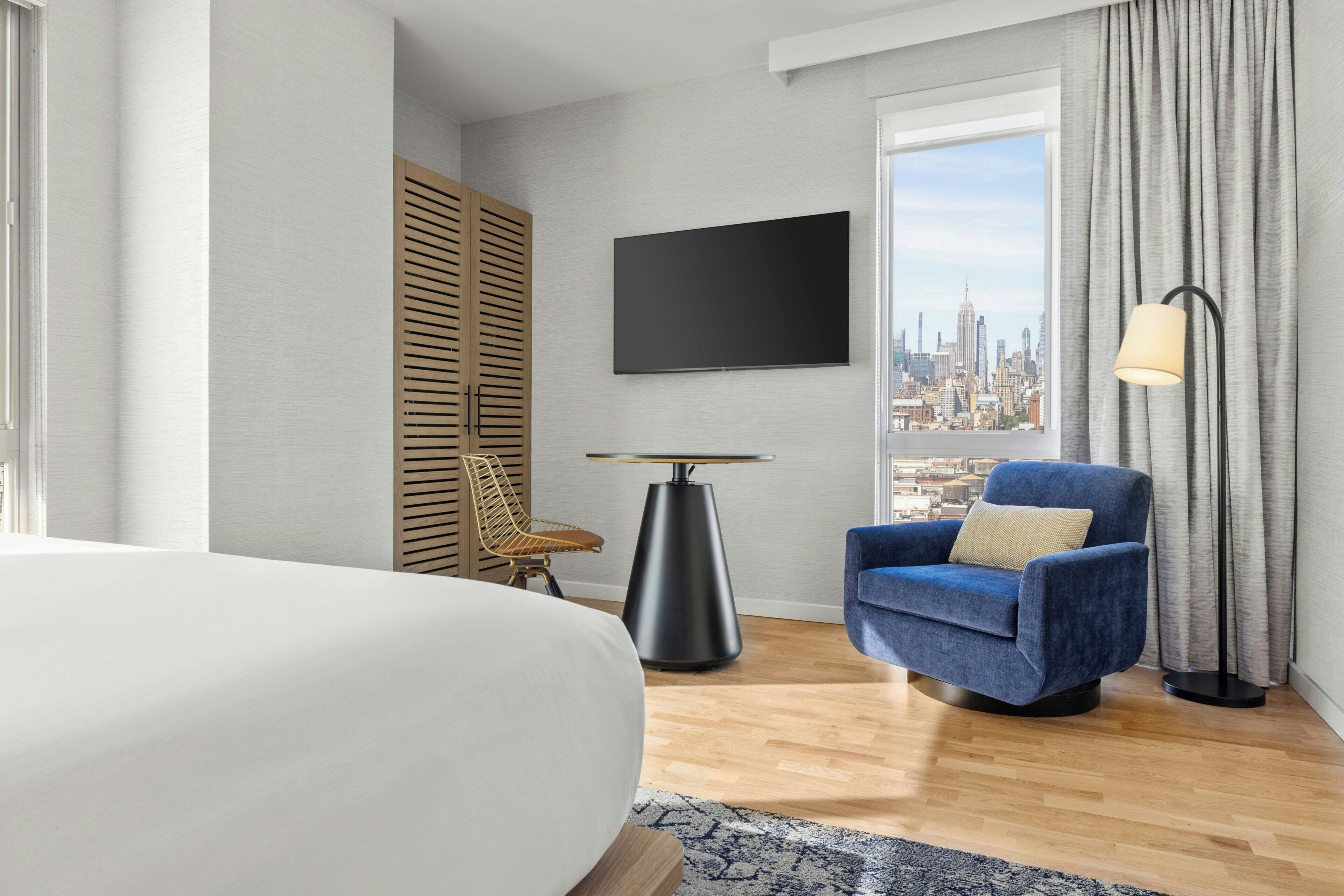 A hotel room with a bed, a chair, a TV, a table, and a view of a cityscape.