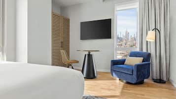 A hotel room with a bed, a chair, a TV, a table, and a view of a cityscape.