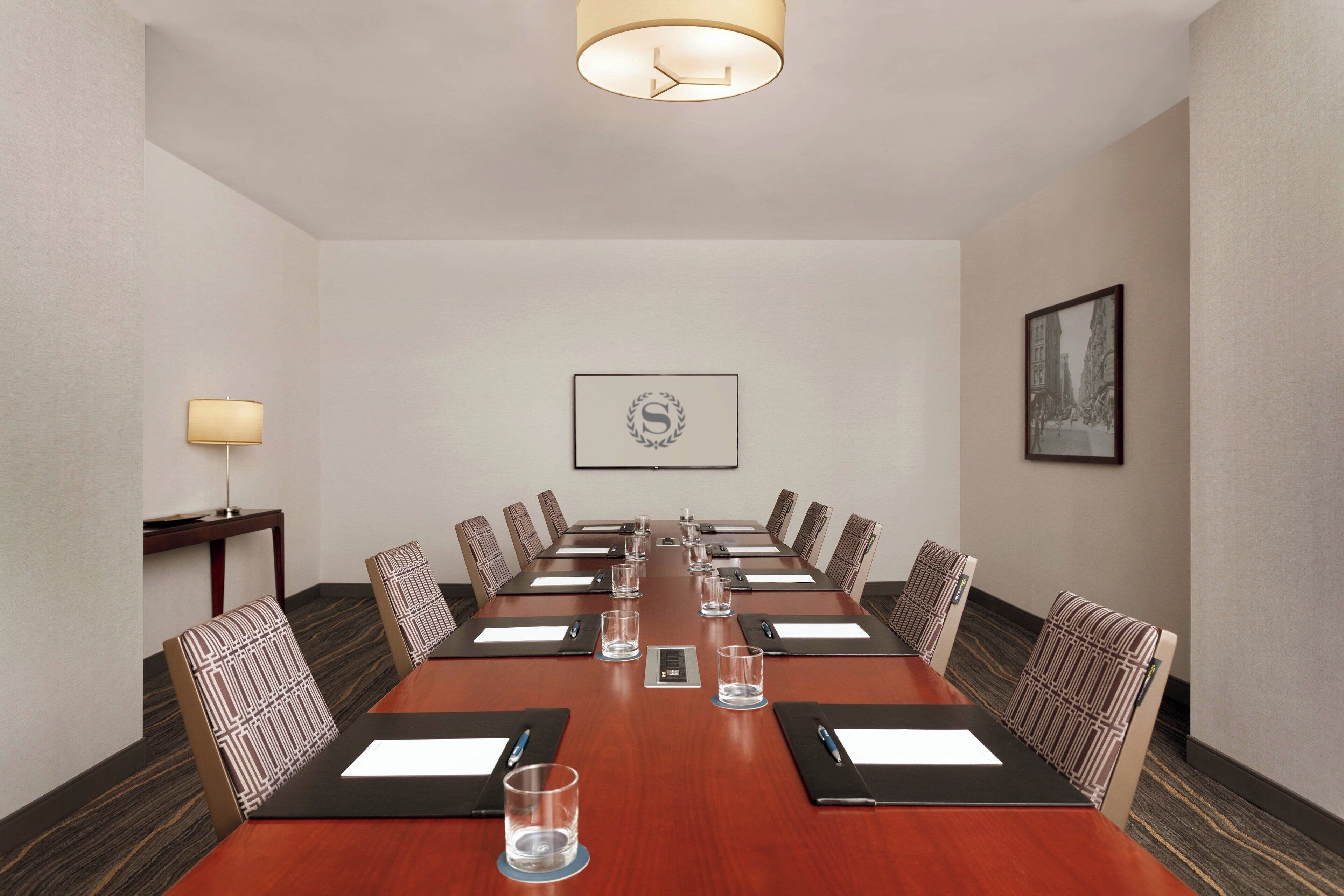 Un salón de conferencias con una mesa larga, sillas, una pantalla de proyector y un cuadro en la pared.