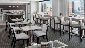 Een moderne hotel eetruimte met hoge tafels en stoelen, een keuken op de achtergrond en een stadszicht door grote ramen.