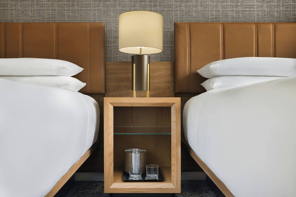 Een hotelkamer met twee bedden, een nachtkastje met een lamp en een klein plankje met een bakje en glazen.