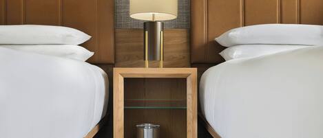 Habitación de hotel con dos camas, una mesita de noche con lámpara y un estante pequeño con un recipiente y vasos.