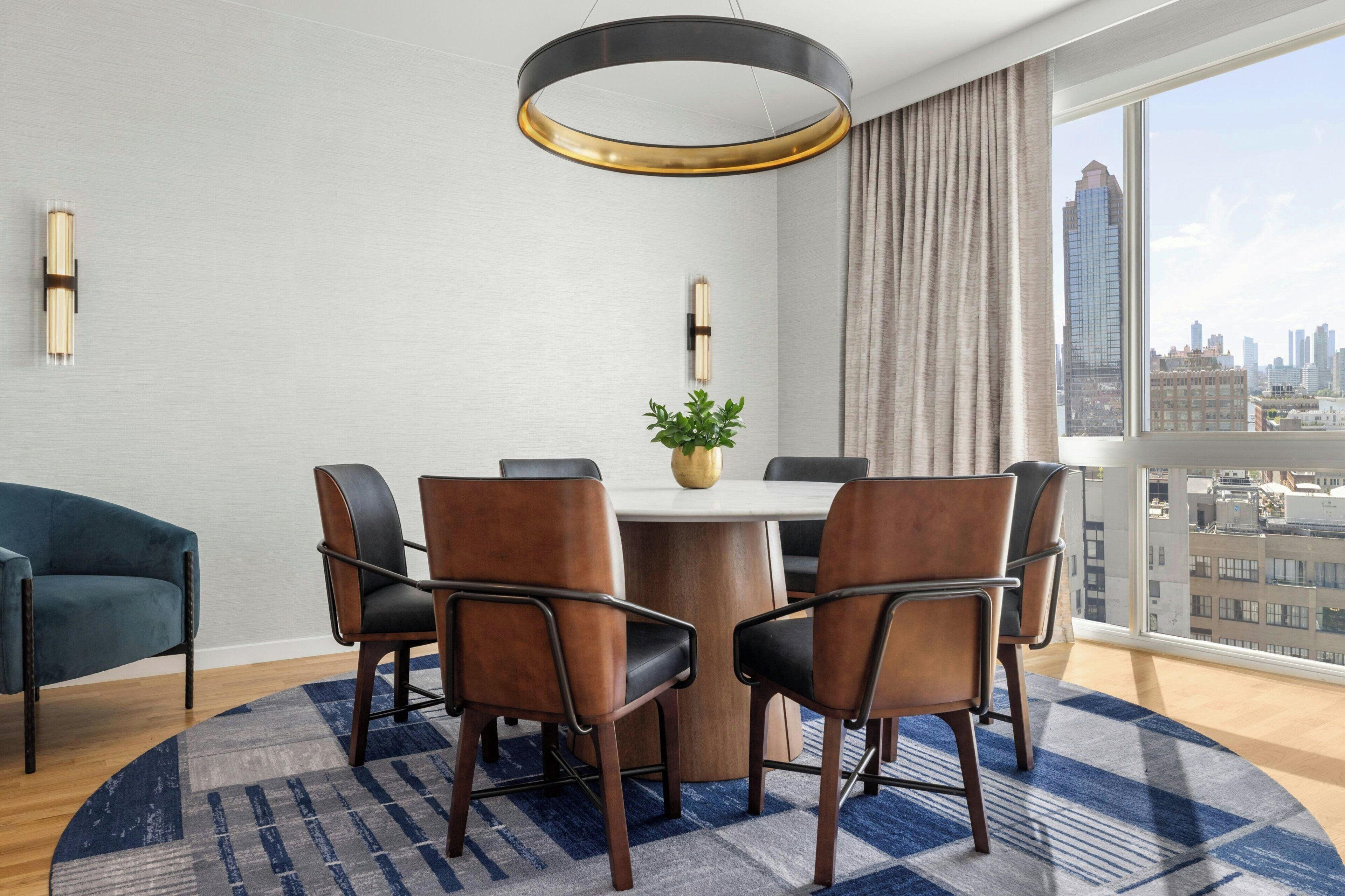 Un comedor moderno con una mesa redonda, seis sillas, una planta en maceta y vistas a un paisaje urbano a través de ventanas amplias.