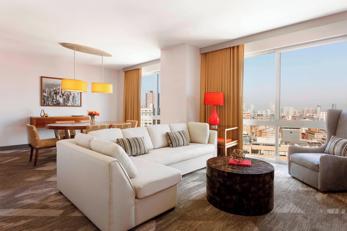 Une chambre d’hôtel moderne dotée d’une grande fenêtre donnant sur la ville, d’un canapé, d’un fauteuil, d’une table à manger et d’une lampe rouge.