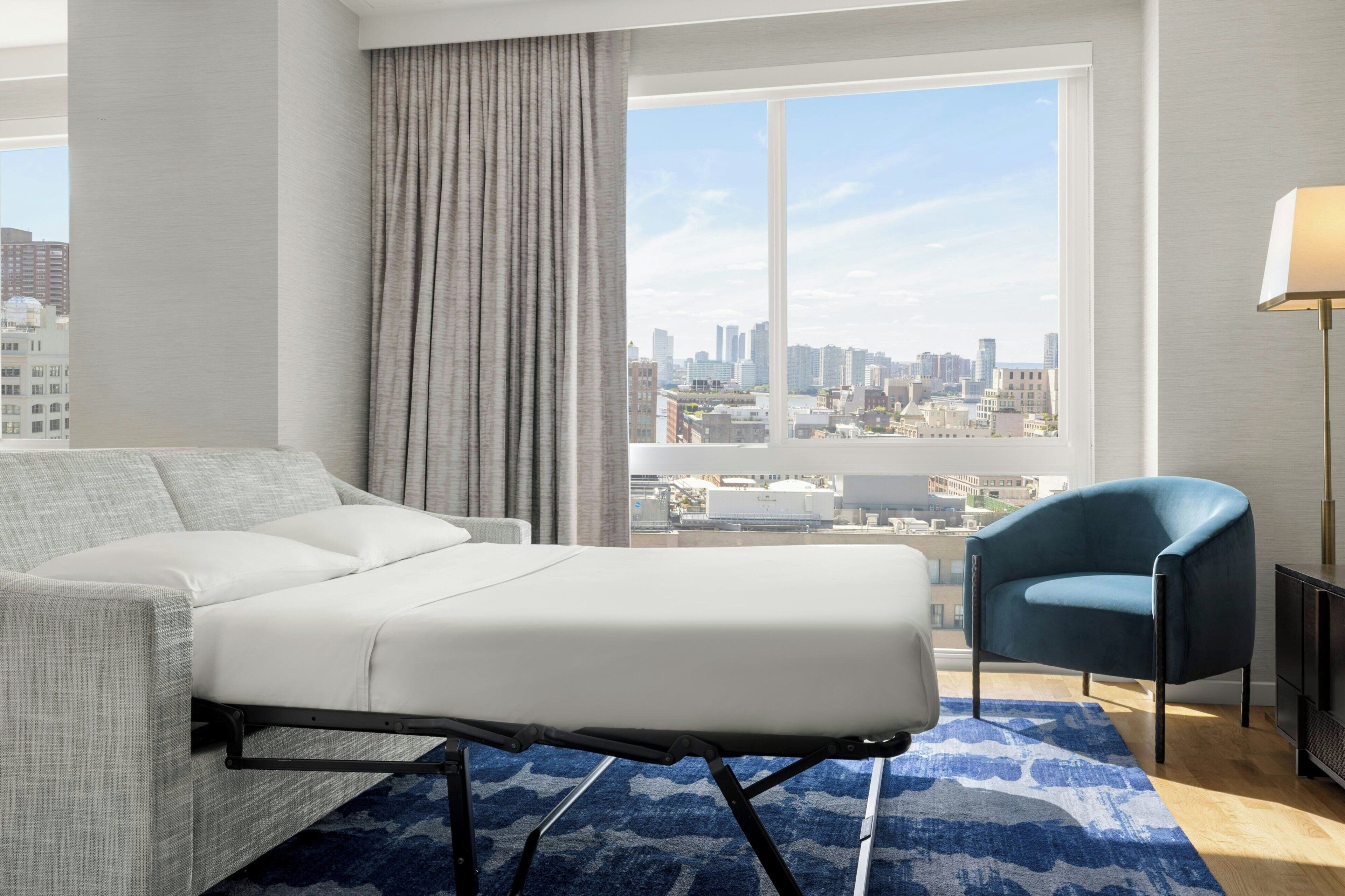 A modern hotel room with a city view, a grey sofa, a blue armchair, and a patterned rug.