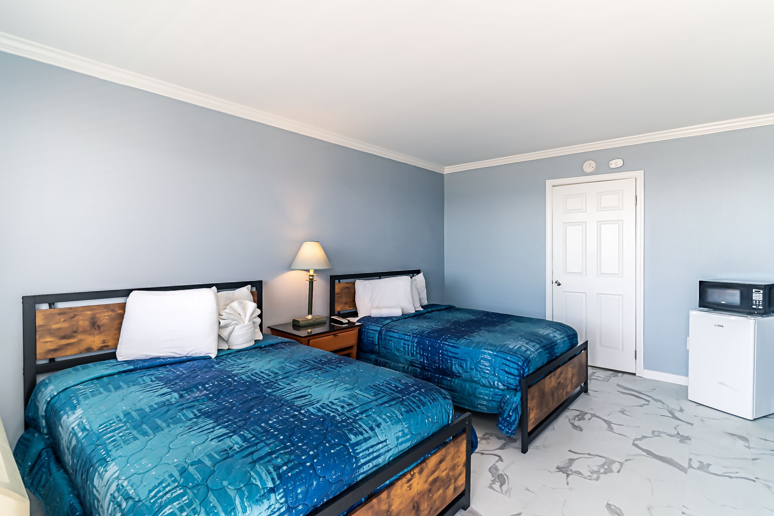 A bedroom with two beds, a nightstand, a lamp, a door, and a microwave.