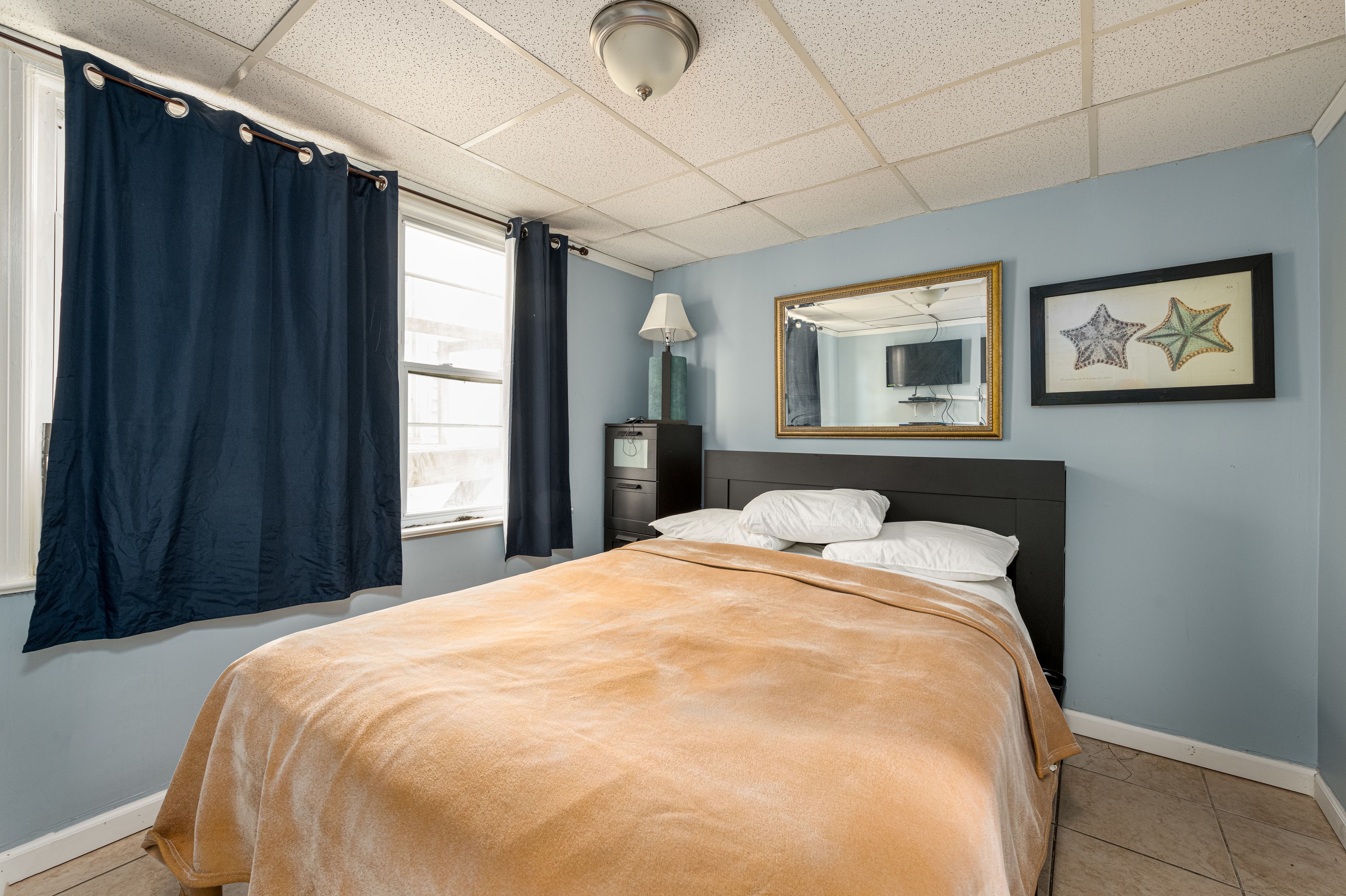 A bedroom with a bed, a window with curtains, a nightstand, a lamp, a mirror, and two framed star decorations on the wall.