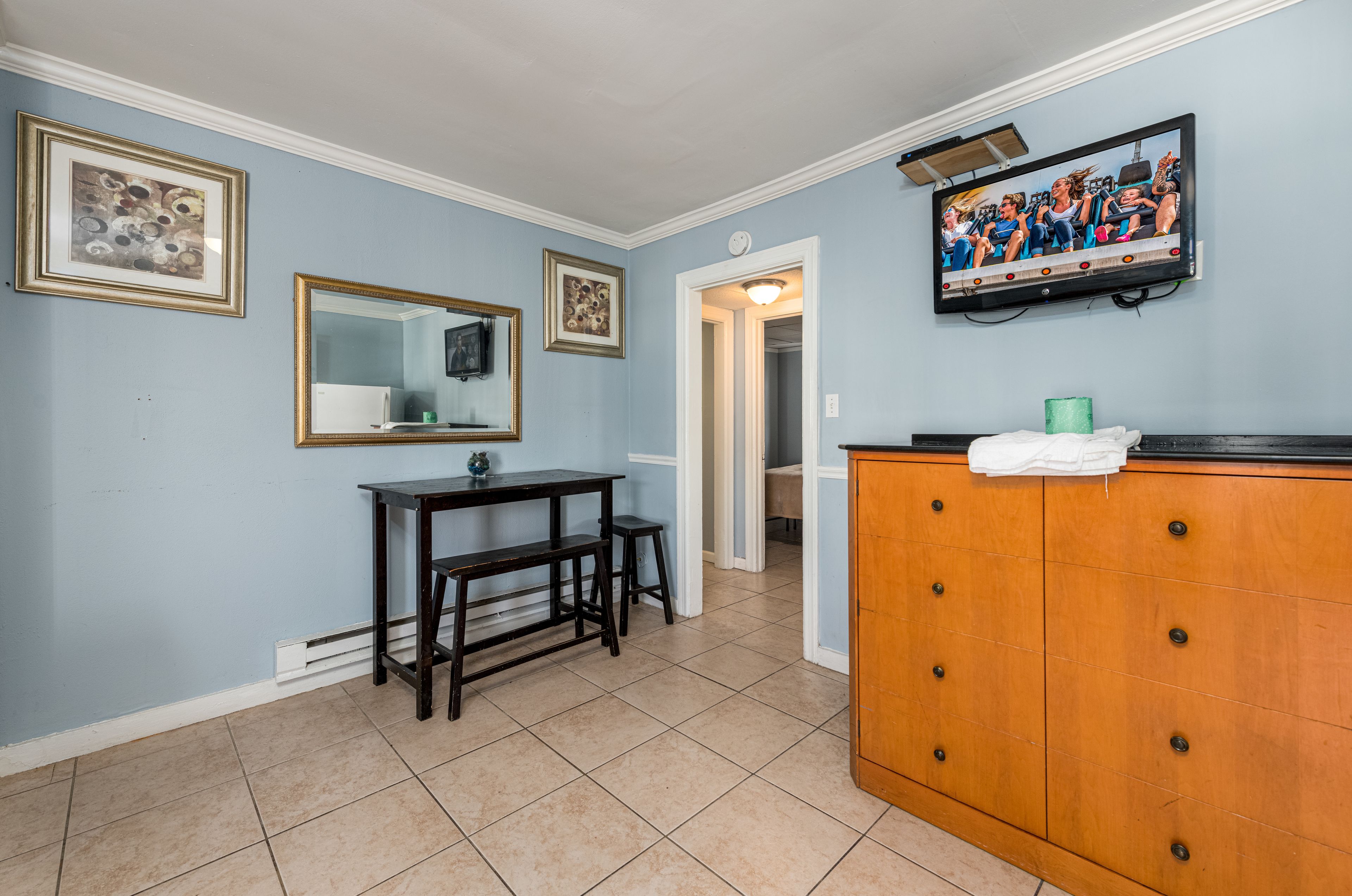 A room with a wooden cabinet, a small table with two stools, a mirror, and a television mounted on the wall.